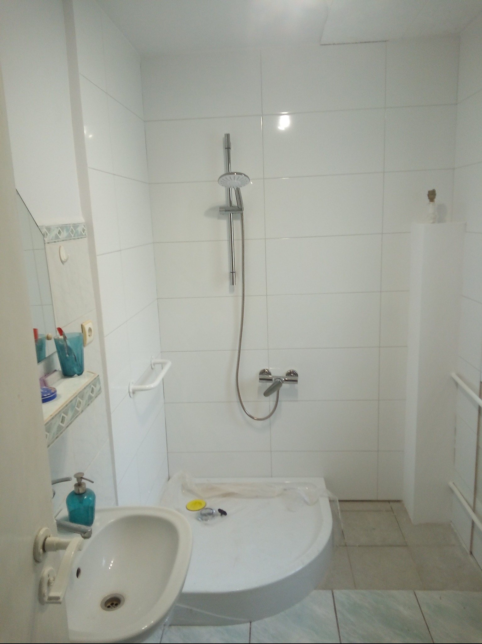 Jasna łazienka z białą armaturą i płytkami. Umywalka z dozownikiem mydła, kabina prysznicowa z deszczownicą. Widoczne akcesoria łazienkowe i fragment lustra.