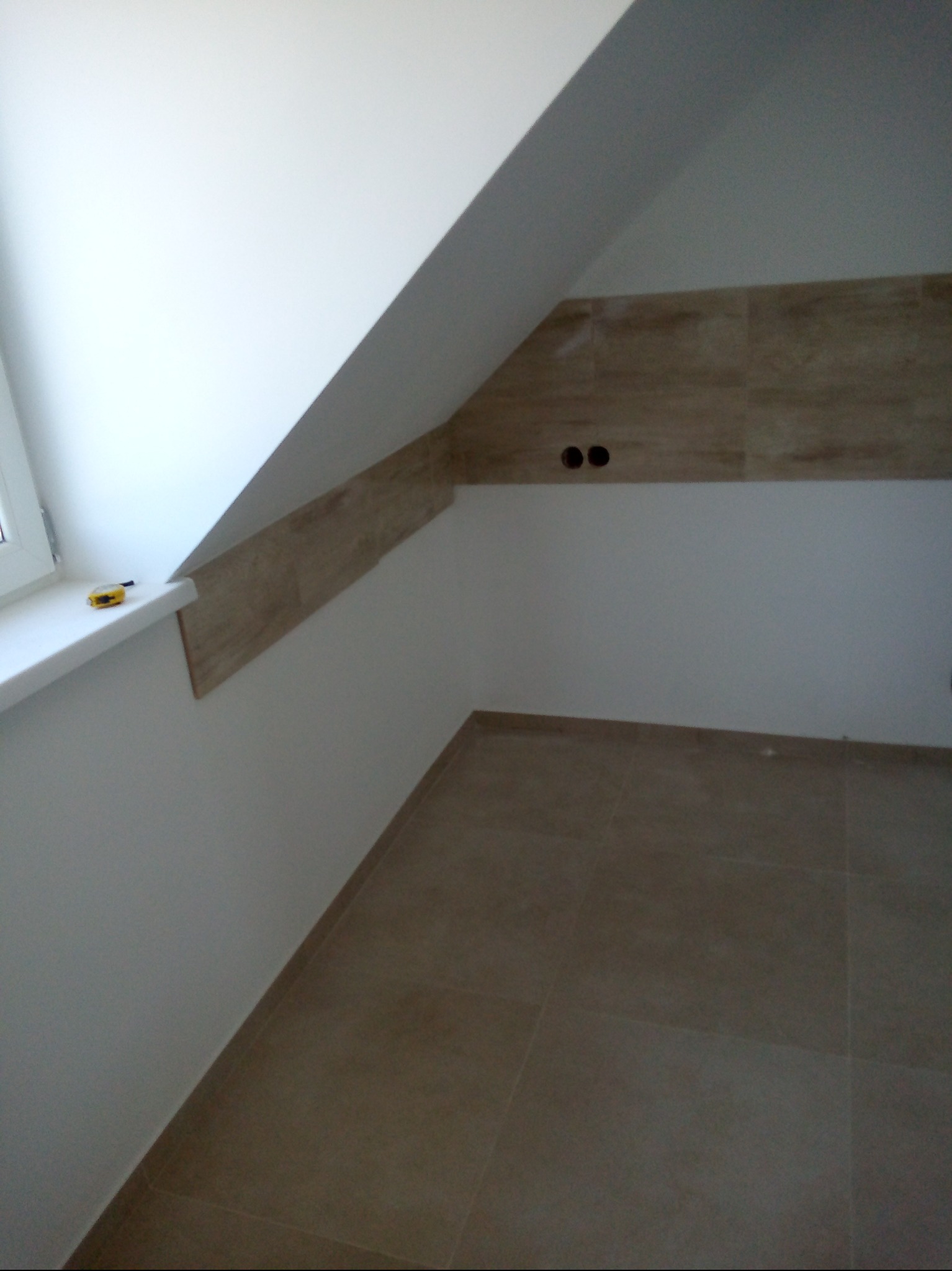 Wykończone poddasze z beżowymi płytkami na podłodze i części ściany, widoczne gniazdka elektryczne i okno z żółtą miarką.