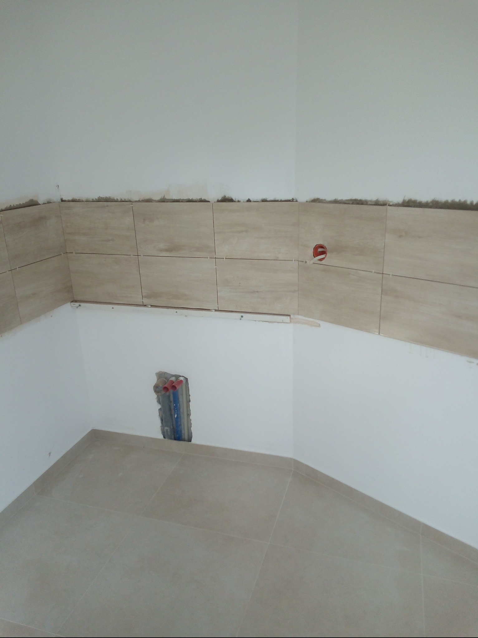 Układanie beżowych płytek na ścianie i podłodze w rogu pomieszczenia. Widoczne wystające rury instalacji wodnej. Etap prac wykończeniowych w łazience.