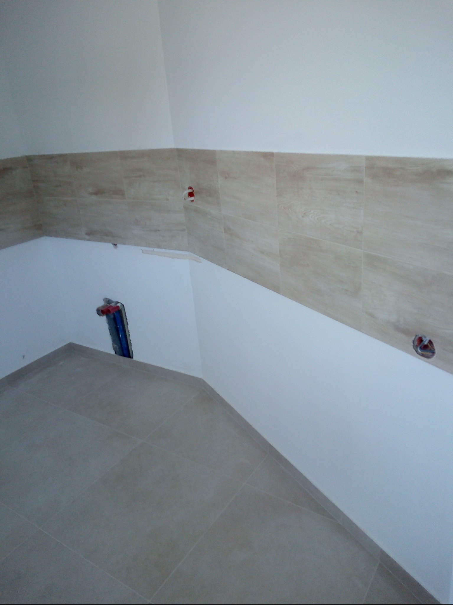 Wykończona łazienka z beżowymi płytkami imitującymi drewno na ścianie i szarymi płytkami na podłodze. Widoczne instalacje elektryczne i wodne.