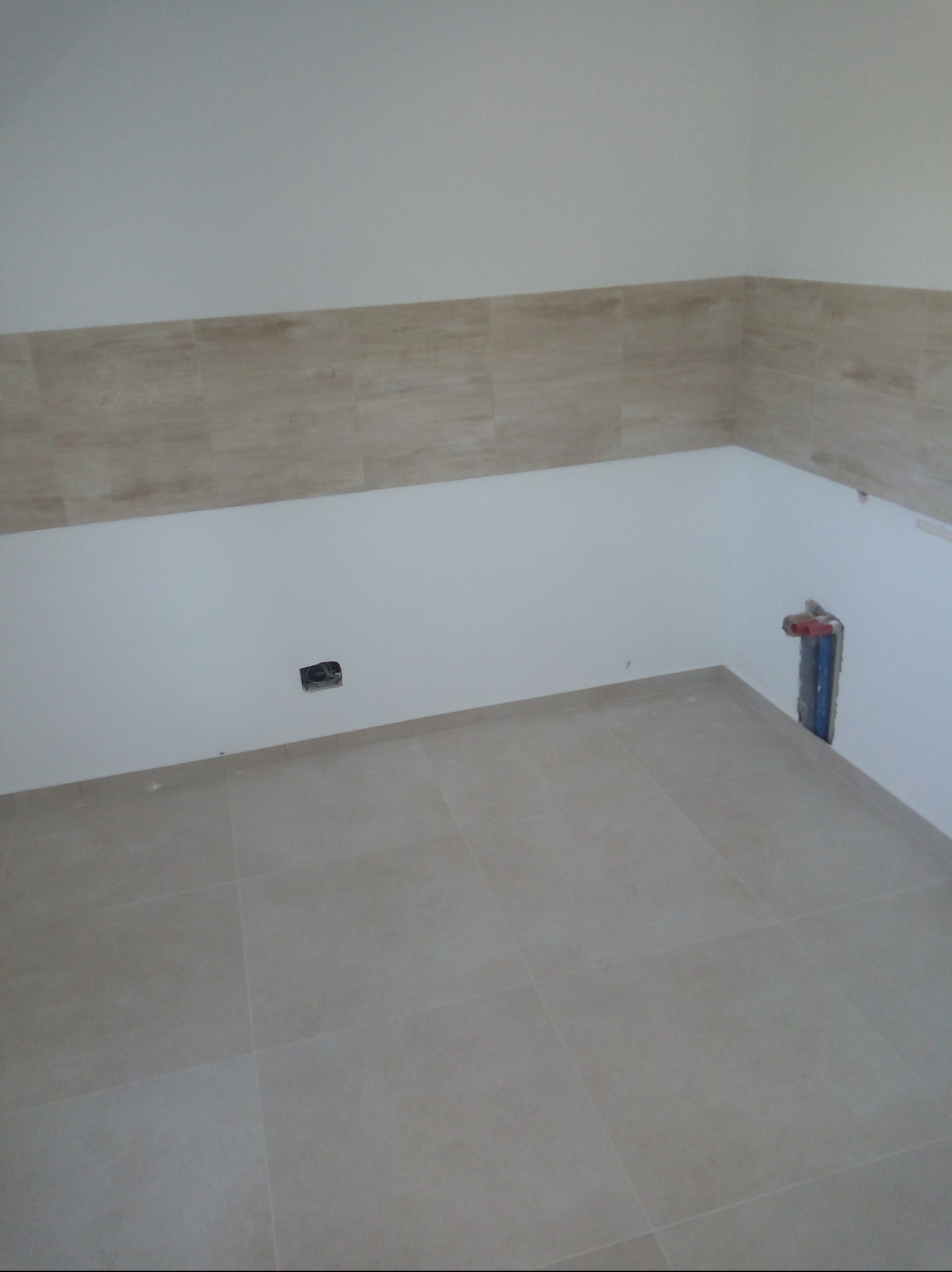 Wykończona łazienka z beżowymi płytkami na podłodze i ścianach do połowy wysokości. Widoczne przyłącza wodne i gniazdko elektryczne. Minimalistyczny design.