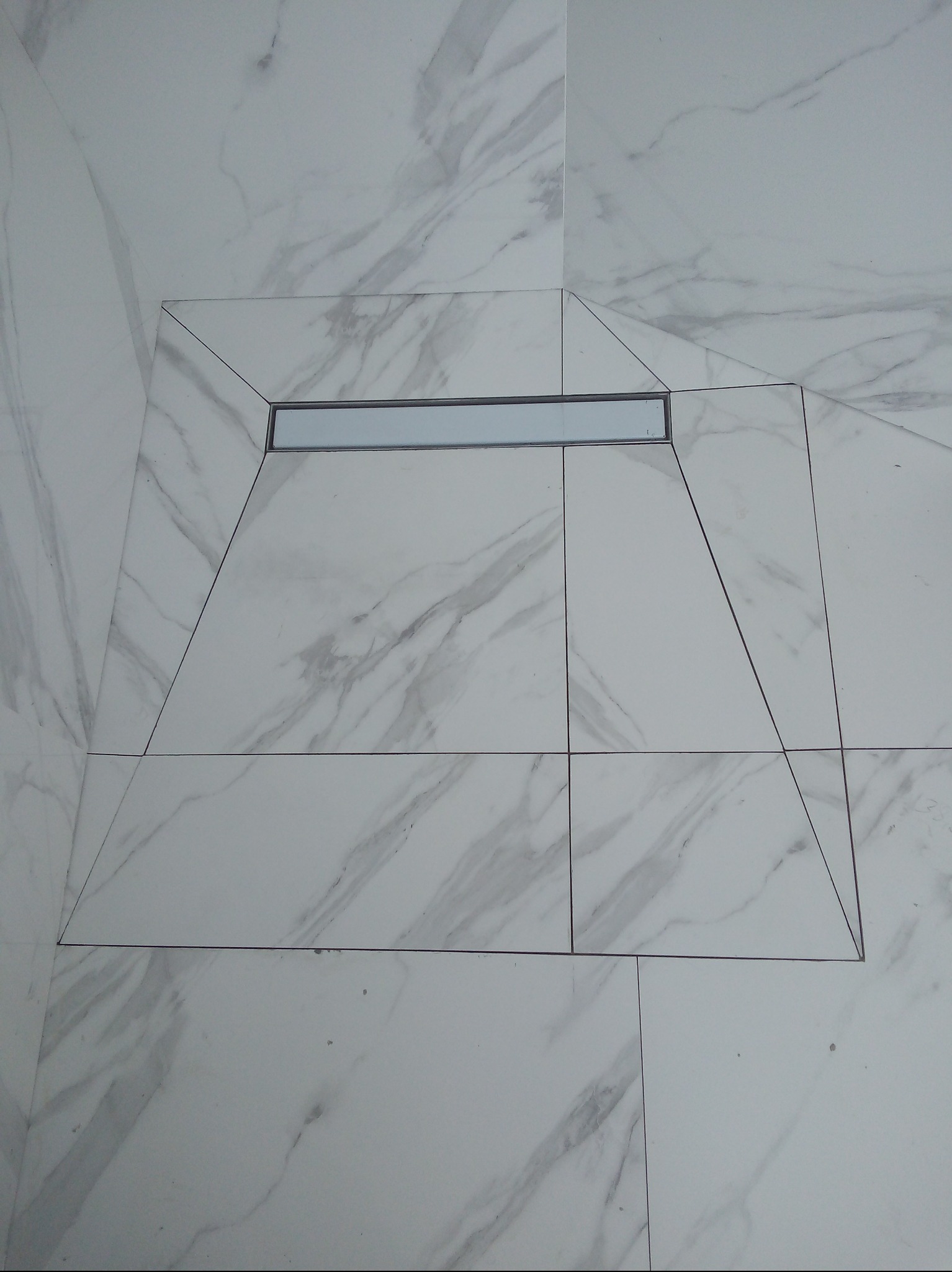 Białe płytki imitujące marmur z odpływem liniowym w kabinie prysznicowej. Precyzyjne cięcia płytek tworzą trapez wokół odpływu. Minimalistyczny design.
