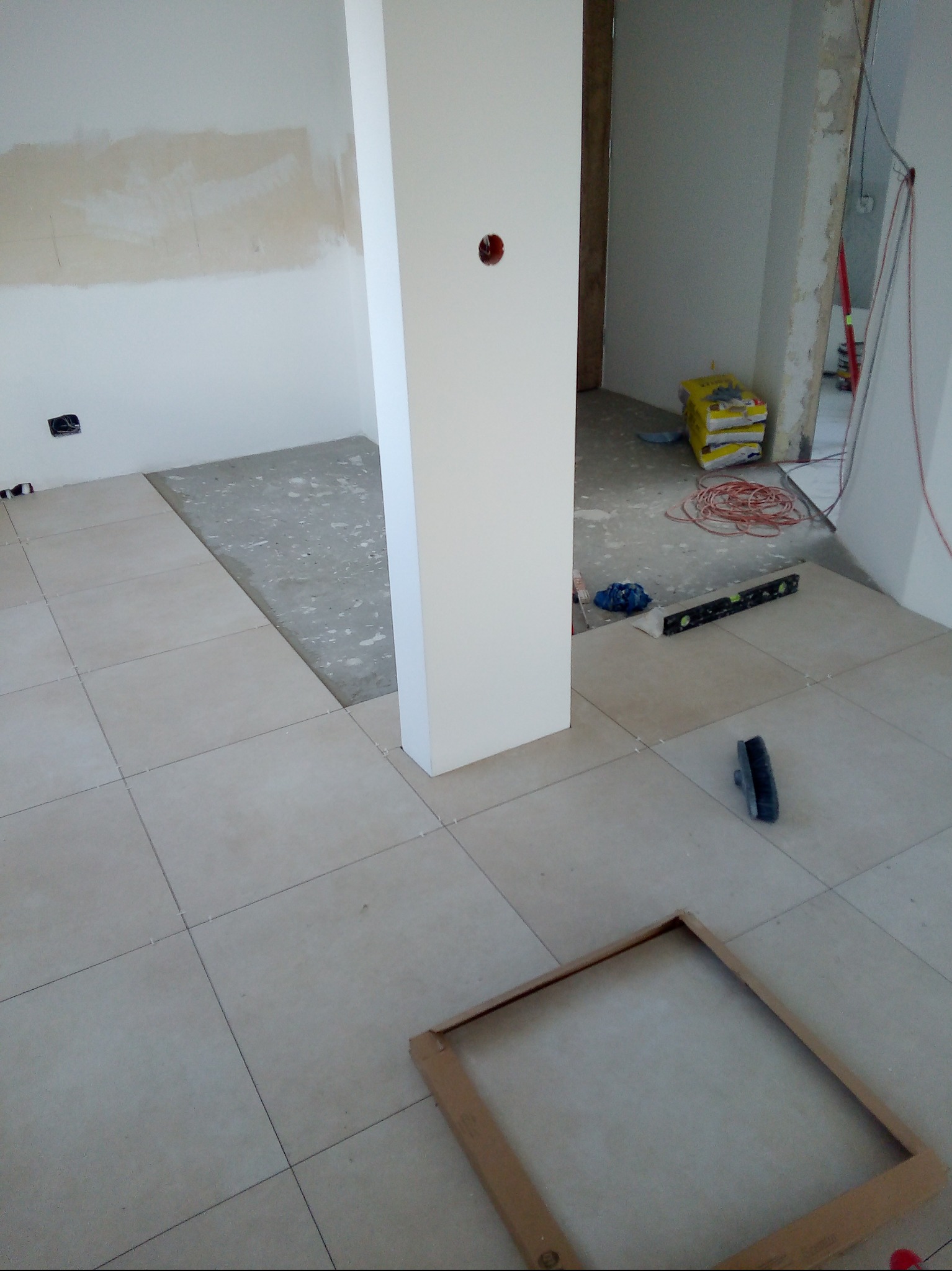 Układanie beżowych płytek podłogowych w pomieszczeniu, widoczny fragment podłogi bez płytek, poziomica, paczki z klejem i drewniana ramka na podłodze.