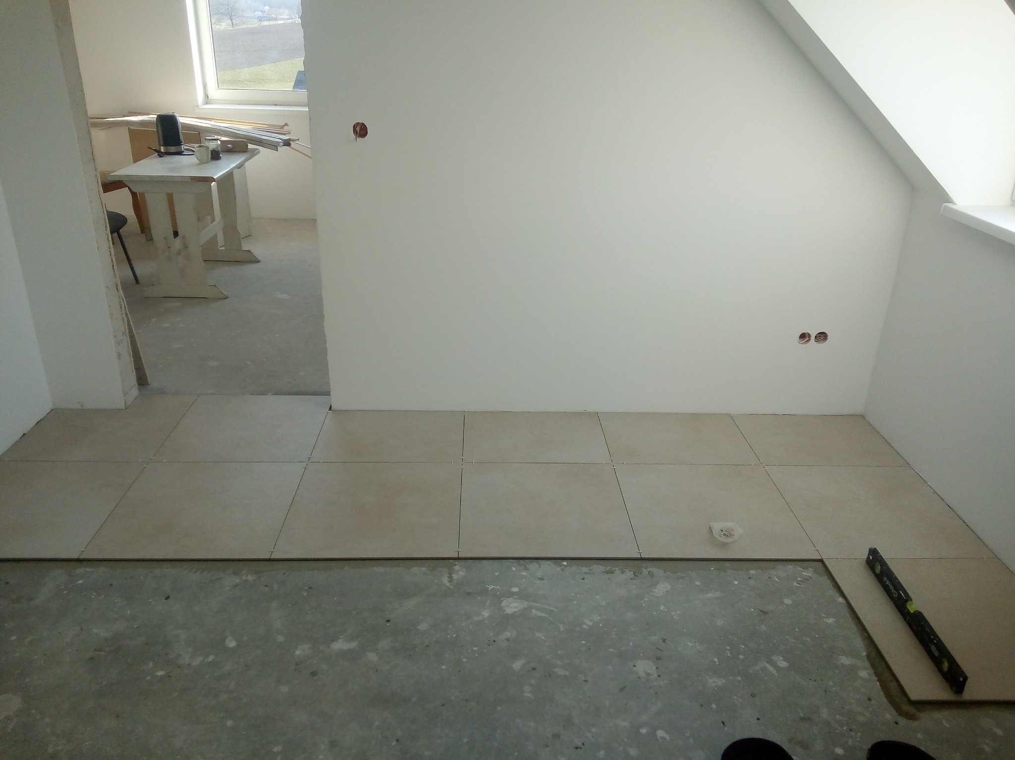 Układanie beżowych płytek podłogowych w jasnym pomieszczeniu na poddaszu. Widoczny poziomica i fragment surowej podłogi przed fugowaniem.