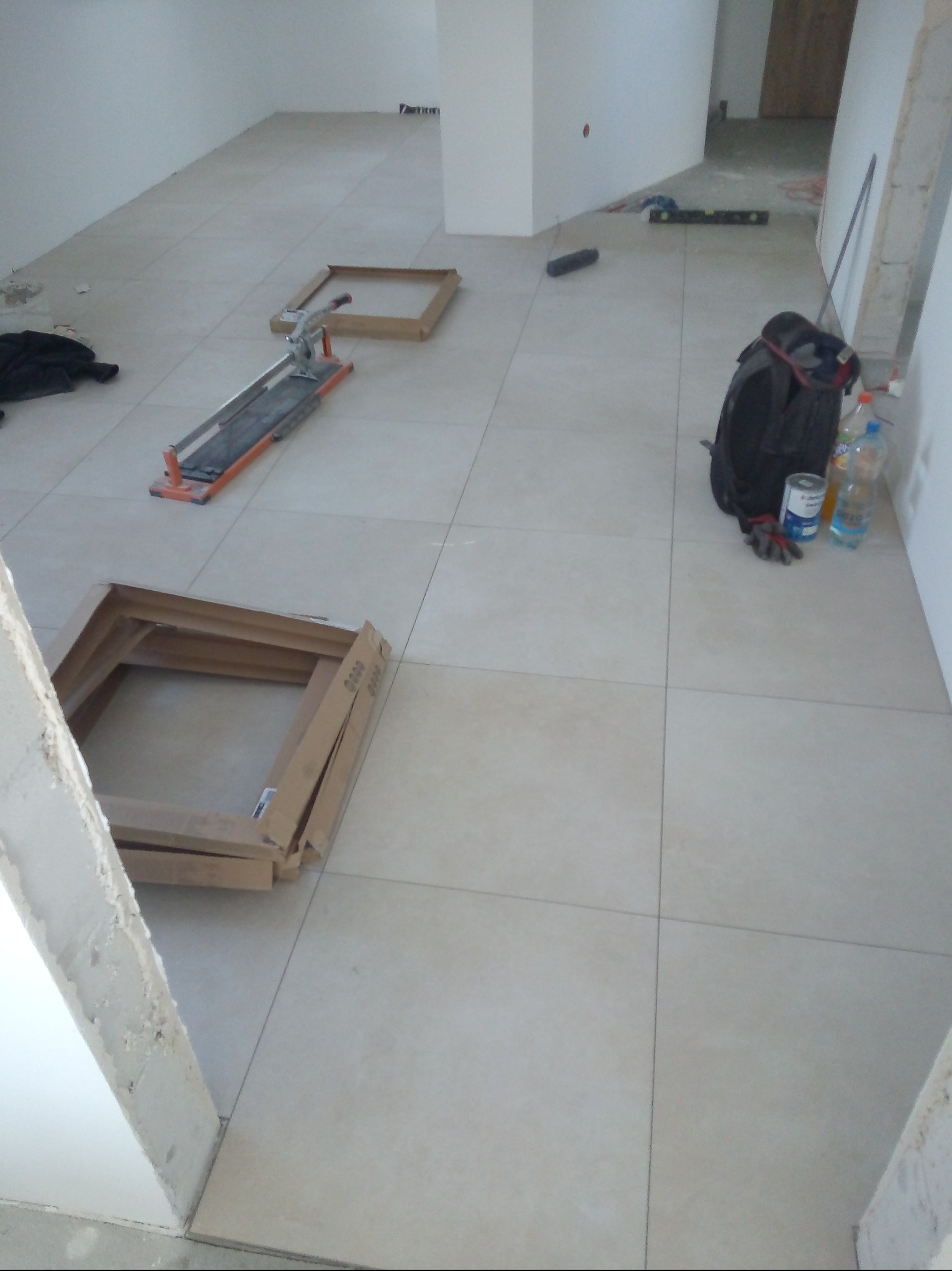 Jasne płytki podłogowe ułożone w nowym pomieszczeniu. Widoczny przecinak do płytek, kartony po płytkach, plecak i poziomica. W tle białe ściany.