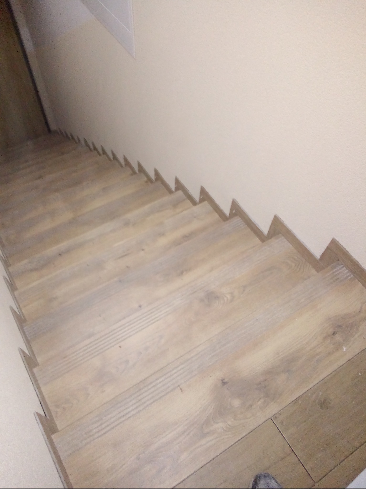 Wykończone schody z panelami w odcieniach beżu i brązu, widok z góry. Ściana w jasnym odcieniu beżu, widoczne listwy przypodłogowe. Układ paneli imituje naturalne drewno.