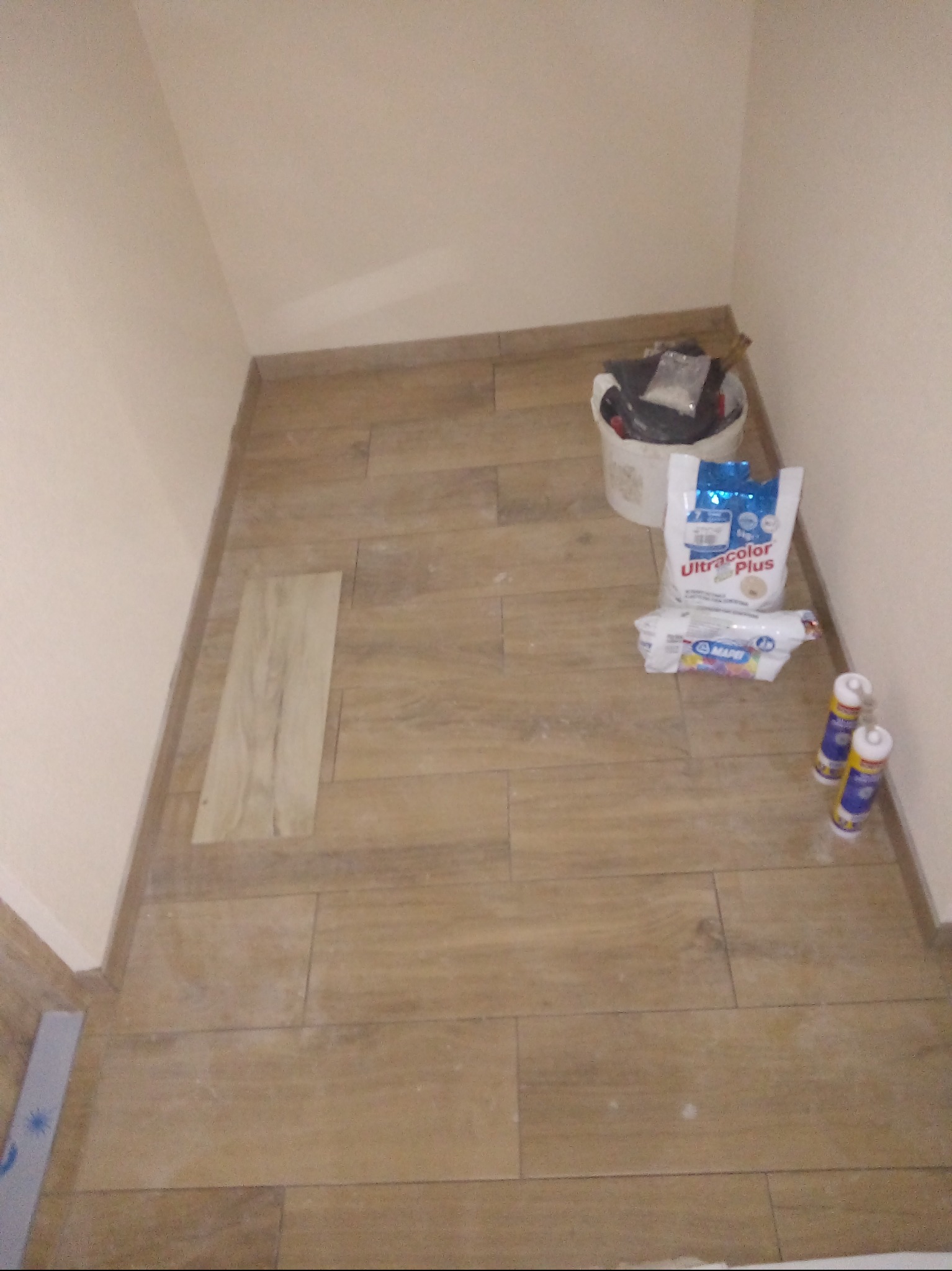 Ułożone płytki podłogowe imitujące drewno w małym pomieszczeniu. Widoczne materiały budowlane: wiadro, worek z zaprawą, fuga i silikon. Jedna płytka nie została jeszcze ułożona.