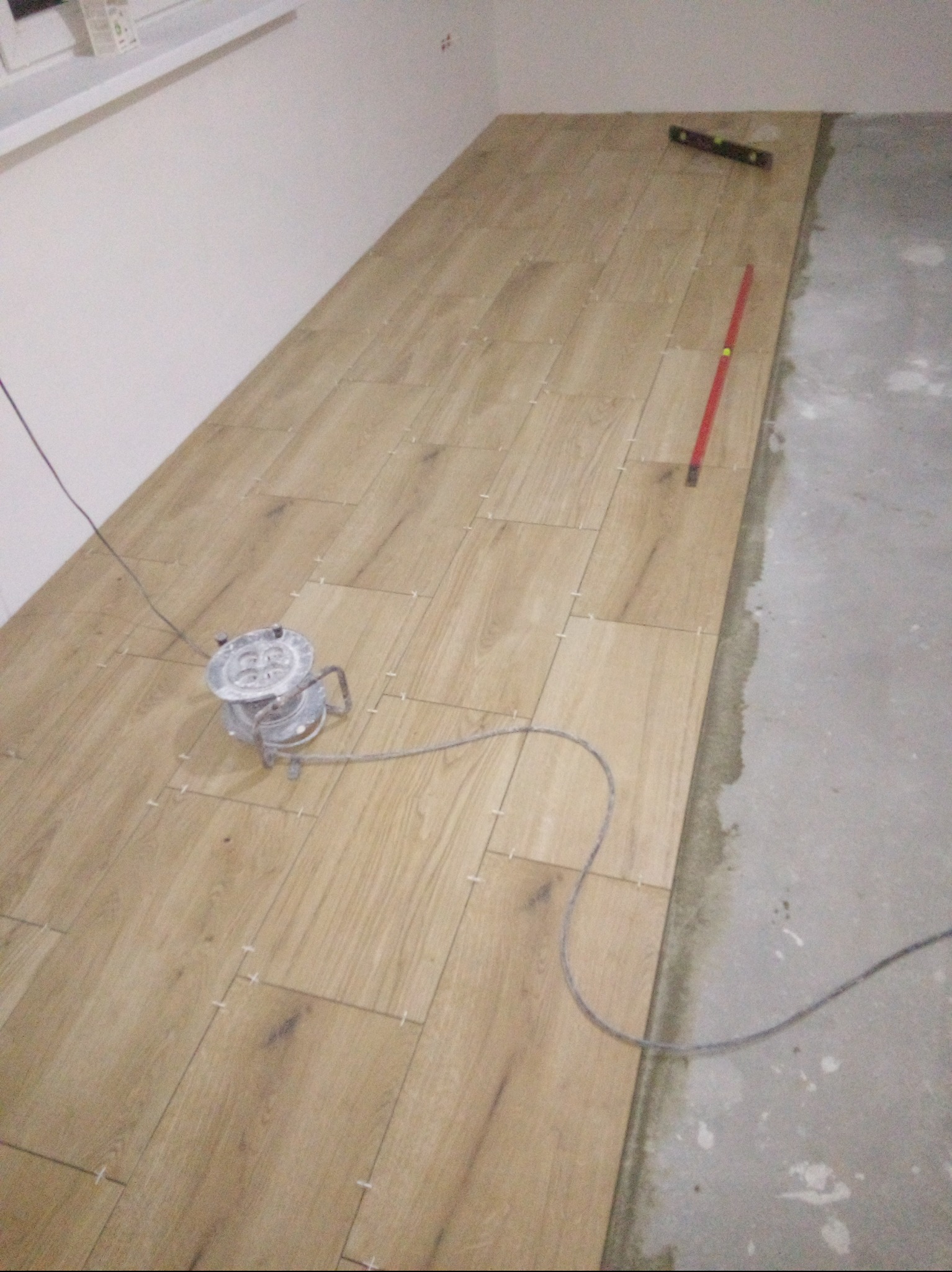 Układanie drewnopodobnych płytek podłogowych z użyciem poziomicy i krzyżyków dystansowych, widoczny kabel zasilający lampę warsztatową.