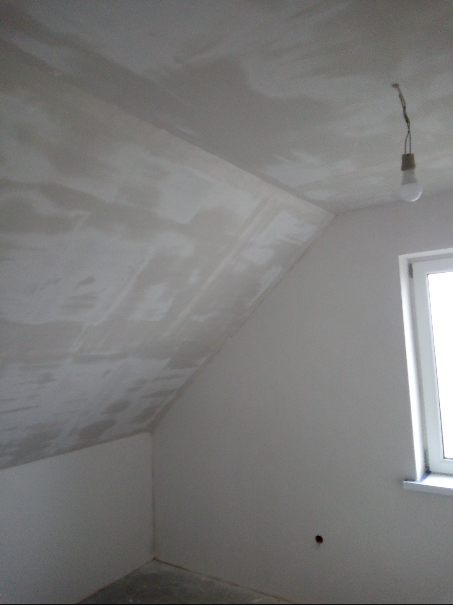 Poddasze ze skośnym sufitem w trakcie prac wykończeniowych. Widoczne ślady po nałożonej gładzi, surowa żarówka i okno. Ściany przygotowane do malowania.