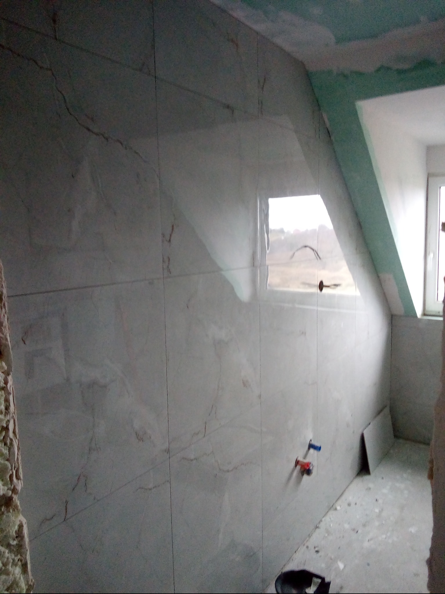 Wykończona łazienka na poddaszu z oknem, ściany wyłożone jasnymi płytkami imitującymi marmur. Widoczne wystające rury i fragment sufitu z płyt karton-gips.