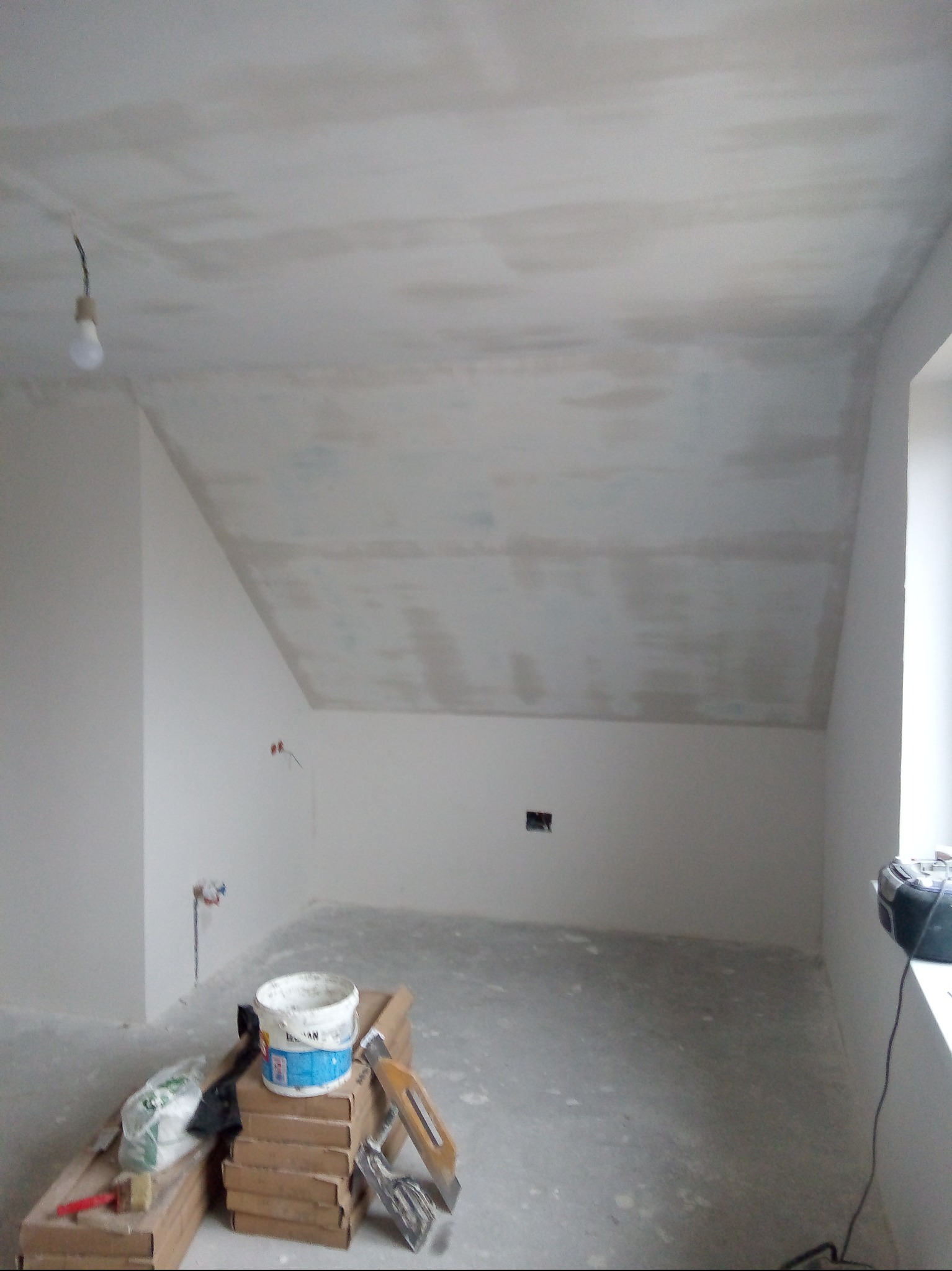 Poddasze w trakcie prac wykończeniowych: ściany i skośny sufit pokryte gładzią, widoczne narzędzia i materiały budowlane. Surowy stan, przygotowanie do malowania.