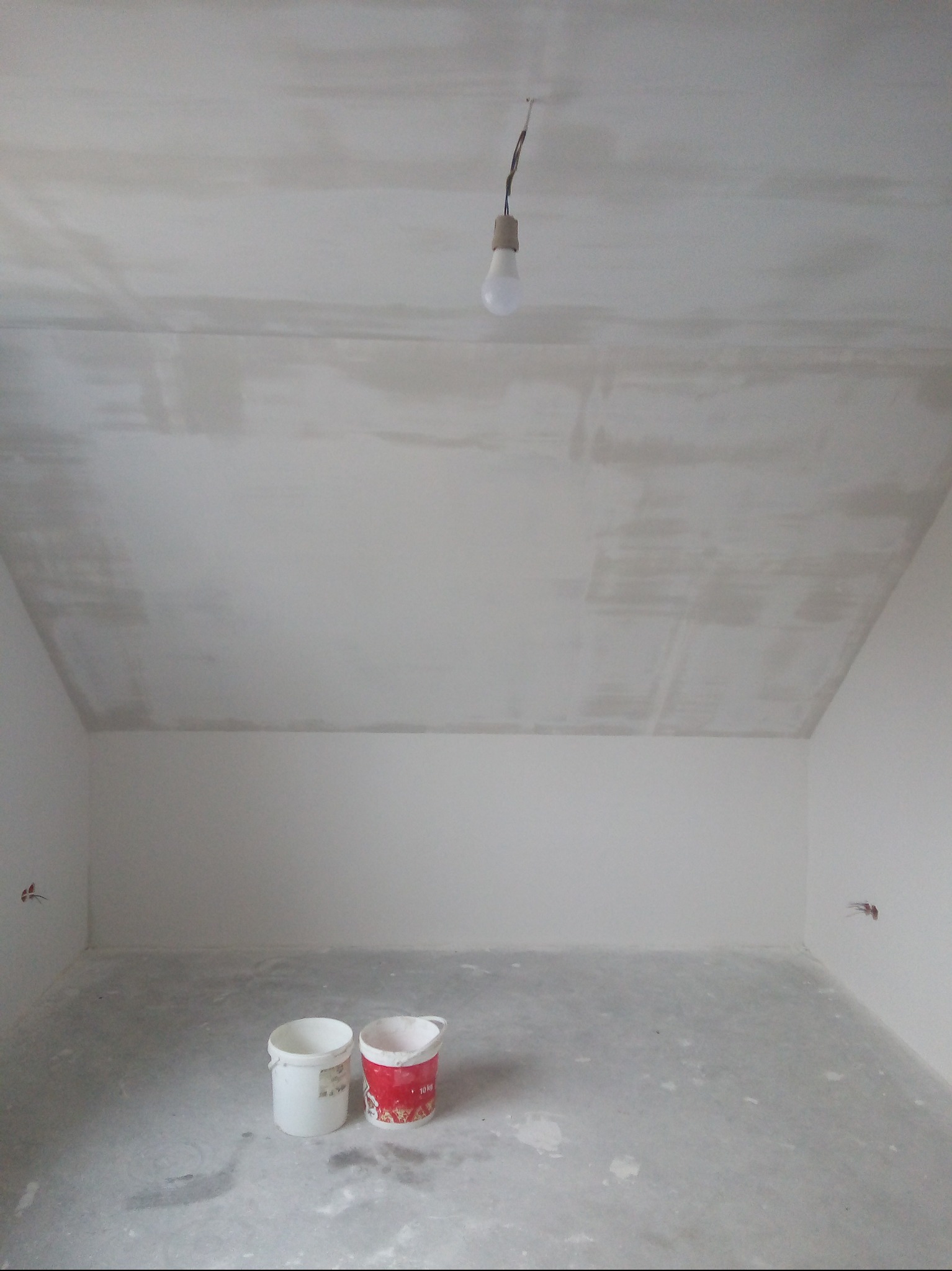 Pokój po gładziowaniu: sufit i ściany pokryte szarą gładzią, na podłodze dwa wiadra po farbie, wisząca żarówka. Widoczne ślady po instalacji elektrycznej.