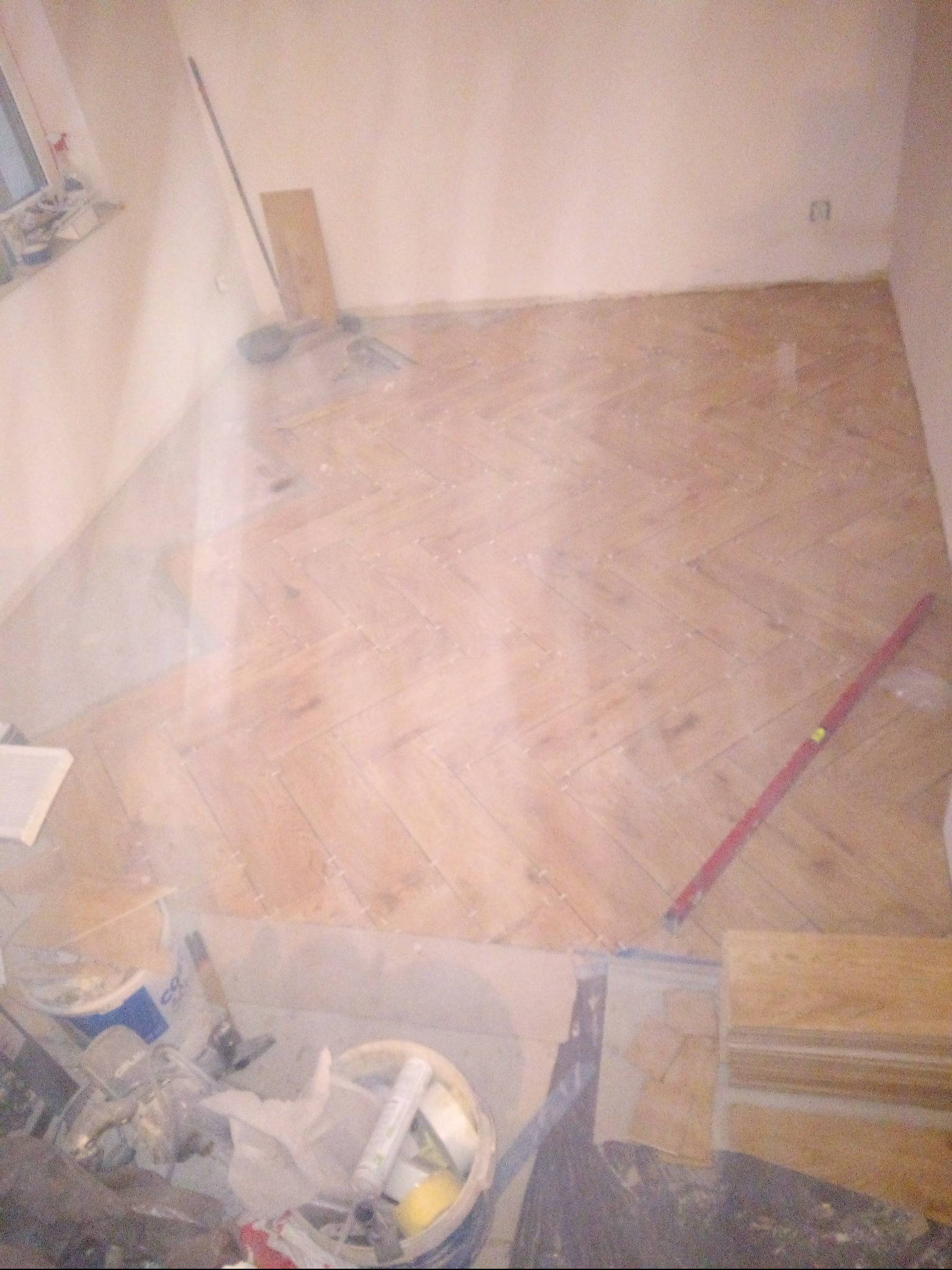 Ułożona podłoga z płytek w jodełkę, widoczny poziomica, wiadro z klejem i inne narzędzia glazurnicze. W tle ściany w trakcie prac wykończeniowych.