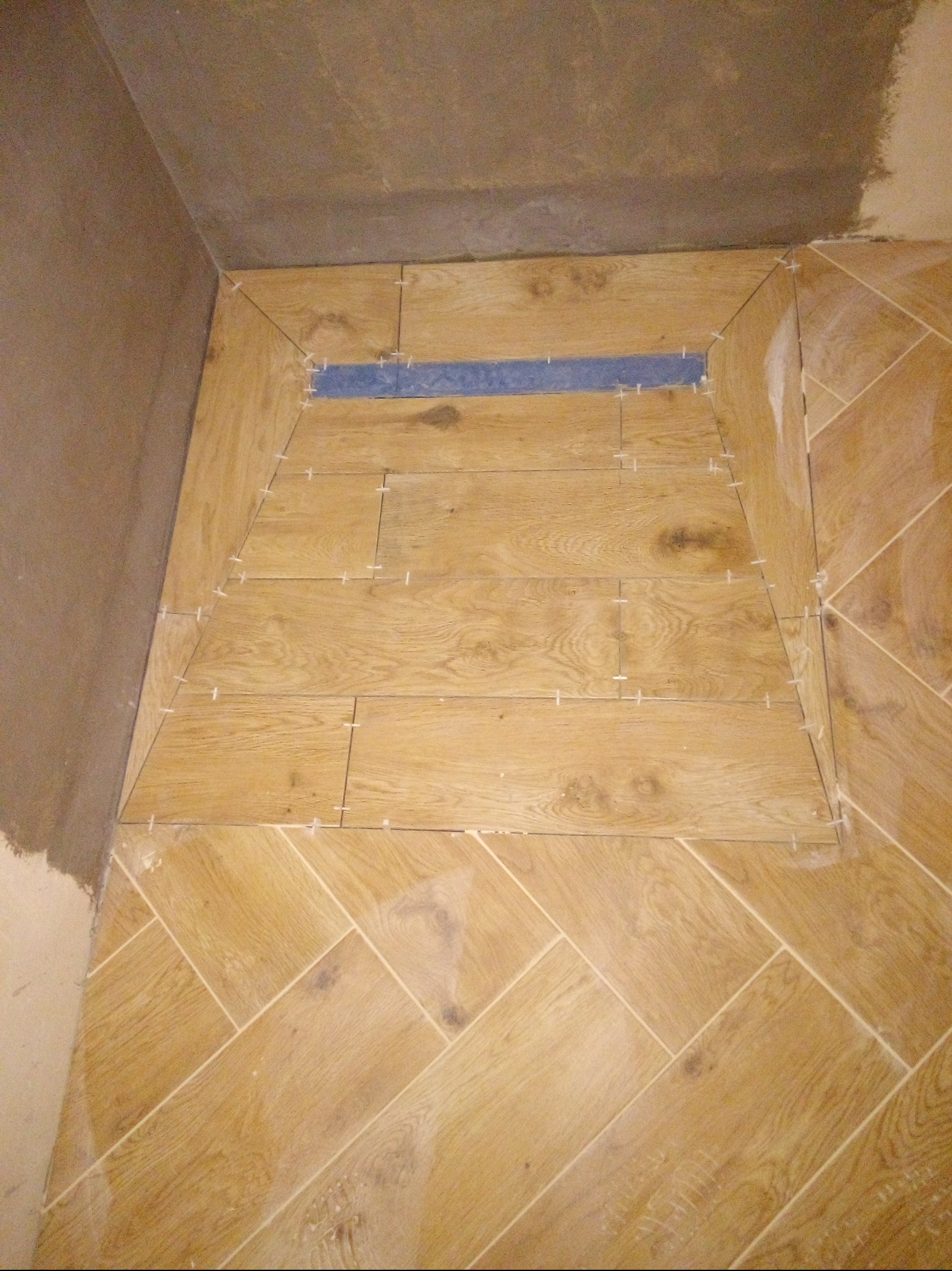 Układanie płytek imitujących drewno w kabinie prysznicowej. Widoczne krzyżyki dystansowe i odpływ liniowy. Fragment ściany z surowym tynkiem.