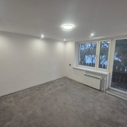 Zgarnę To - Pusty pokój po remoncie: szare panele podłogowe, białe ściany, okno z widokiem na zewnątrz, grzejnik. Minimalistyczne oświetlenie sufitowe.