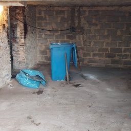 Zgarnę To - Piwnica po sprzątaniu: niebieski kosz na śmieci, worki i narzędzia na betonowej podłodze. Ściany z cegieł i kamienia, surowe oświetlenie.