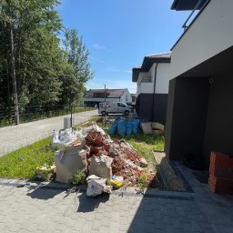 Zgarnę To - Stos gruzu budowlanego z potłuczonych cegieł i worków na trawniku, obok świeżo ułożonej kostki brukowej i nowoczesnych domów szeregowych w słoneczny dzień.