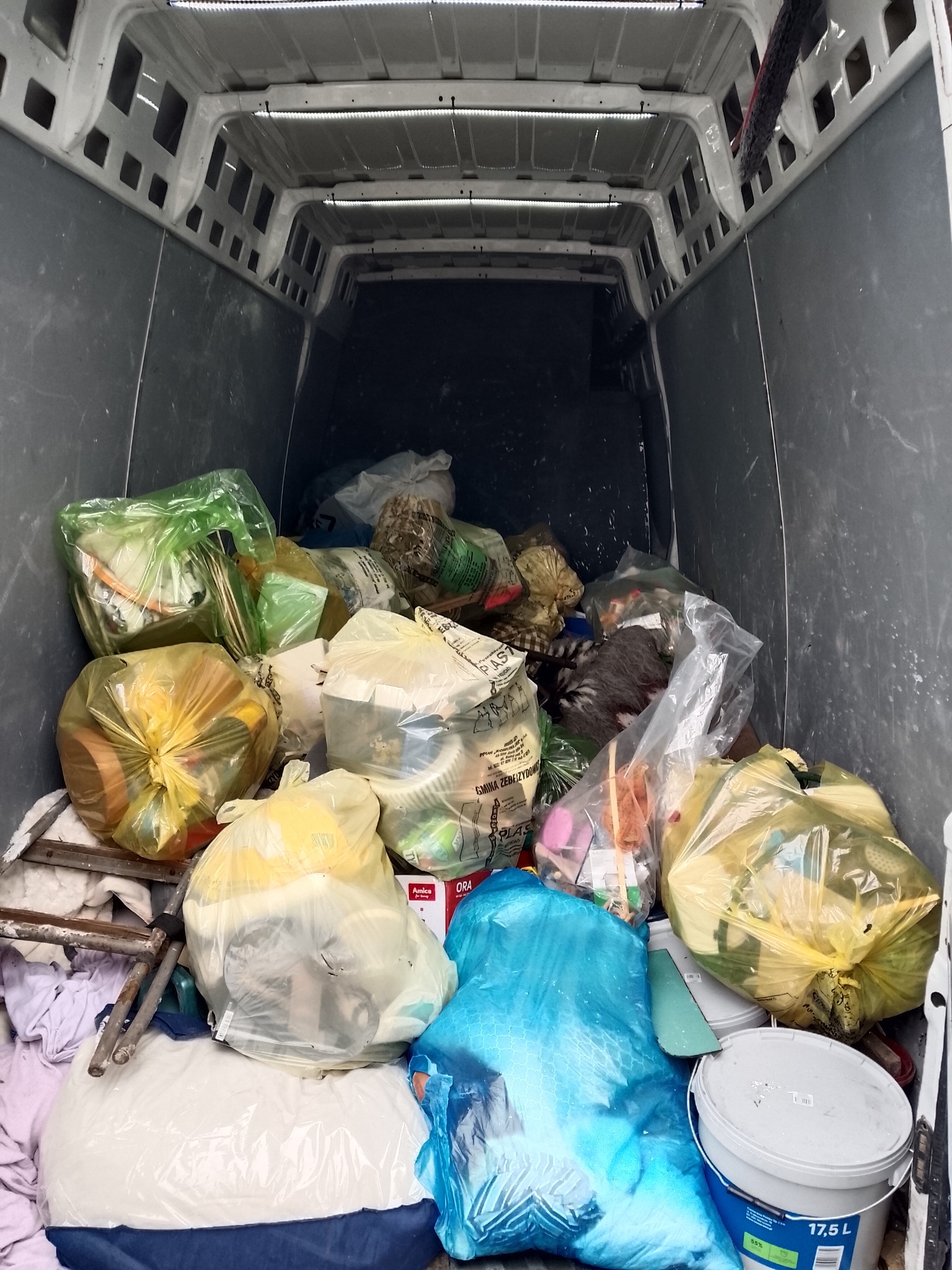 Wnętrze busa wypełnione workami z odpadami, widoczne różne kolory worków i przedmioty wewnątrz, częściowo widoczna drabina i wiadro z farbą.
