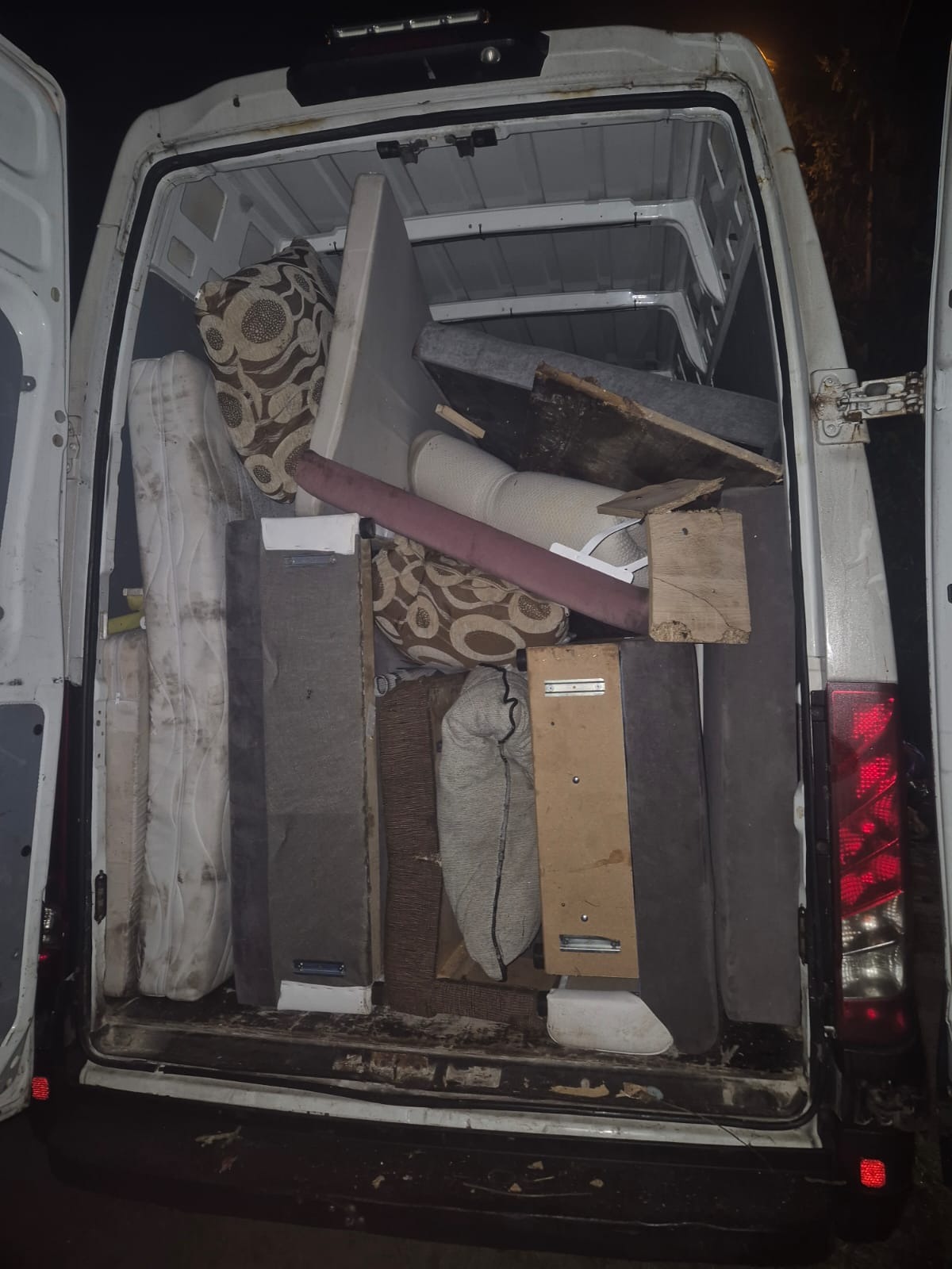 Wnętrze białego busa wypełnione meblami i przedmiotami gospodarstwa domowego, widoczne przez otwarte tylne drzwi. Nocne oświetlenie.