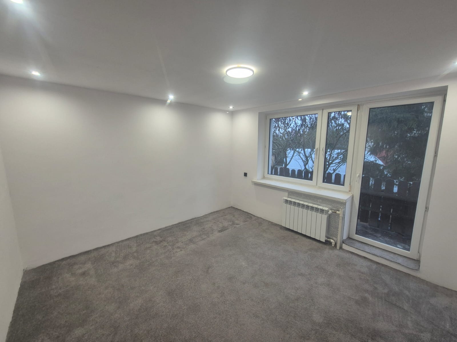 Pusty pokój po remoncie: szare panele podłogowe, białe ściany, okno z widokiem na zewnątrz, grzejnik. Minimalistyczne oświetlenie sufitowe.