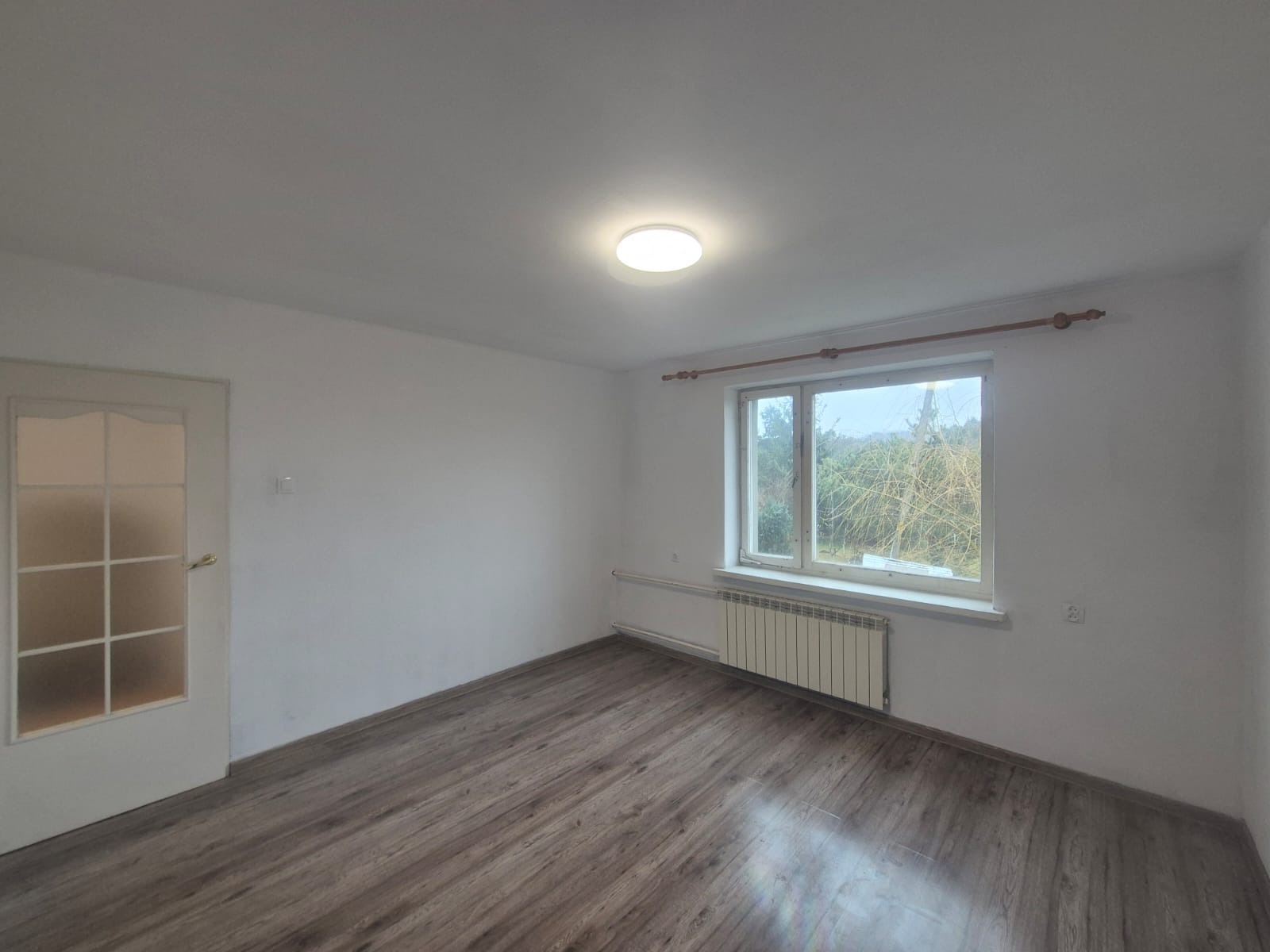 Puste, jasne wnętrze pokoju z panelami podłogowymi, białymi ścianami, oknem z widokiem na drzewa i grzejnikiem. Widoczna biała, przeszklona rama drzwi.
