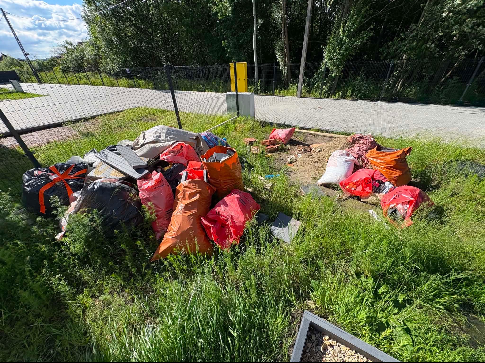 Duża ilość odpadów budowlanych, w tym styropian w pomarańczowych i czerwonych workach, leżąca na trawniku przy chodniku z kostki brukowej, obok czarnego ogrodzenia.