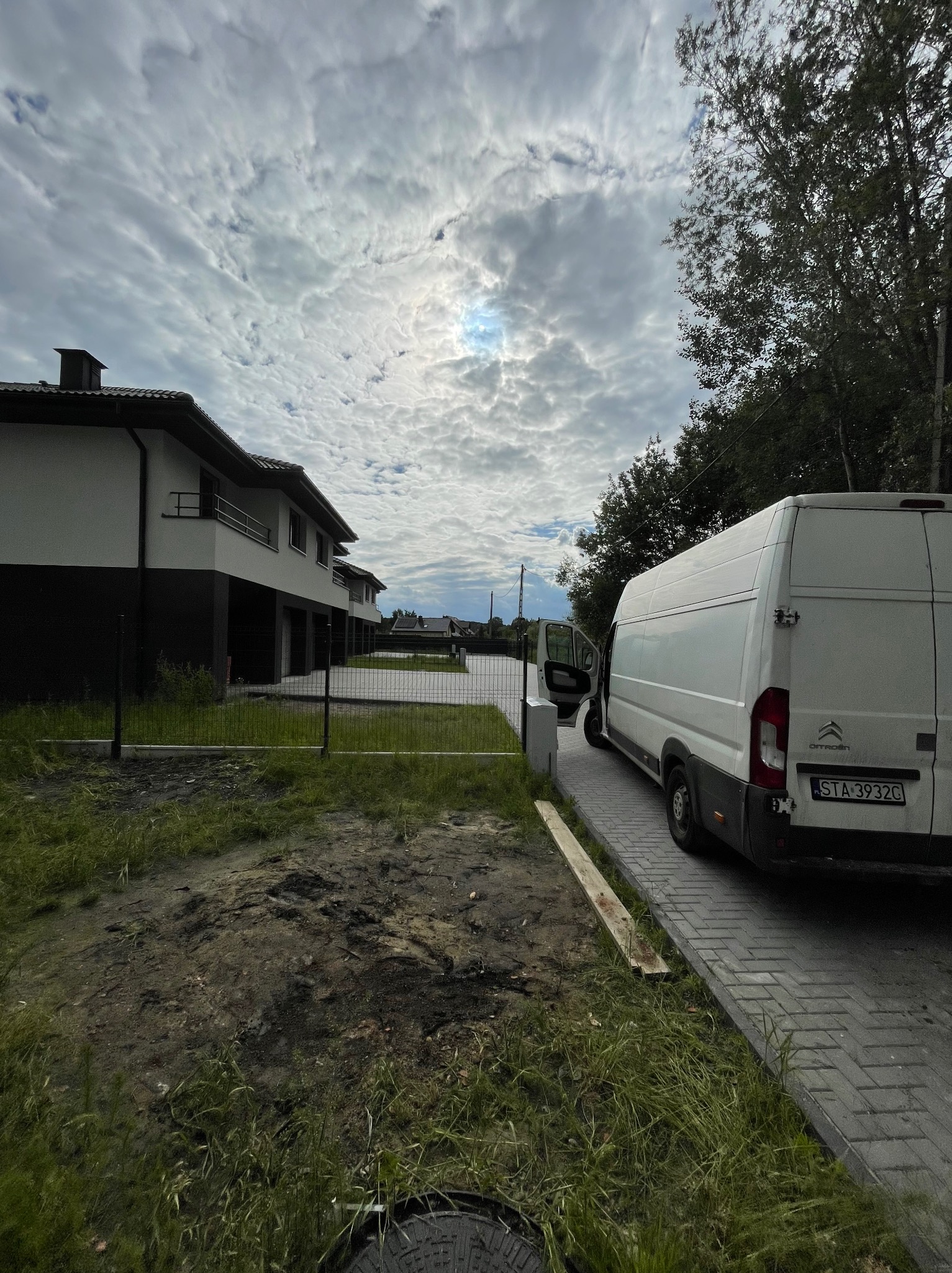 Biały dostawczy Citroen z otwartymi drzwiami bocznymi, zaparkowany na kostce brukowej przed szeregiem nowoczesnych domów. W tle zachmurzone niebo, na pierwszym planie trawnik i ziemia.