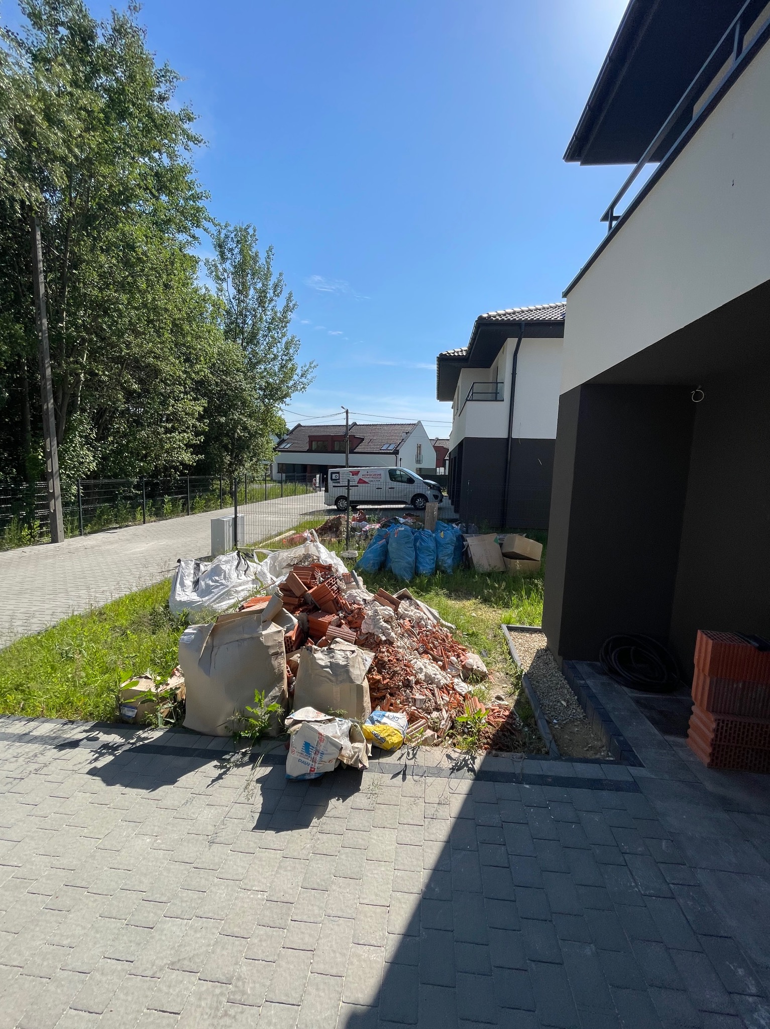 Stos gruzu budowlanego z potłuczonych cegieł i worków na trawniku, obok świeżo ułożonej kostki brukowej i nowoczesnych domów szeregowych w słoneczny dzień.