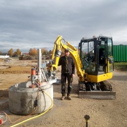 Jakub Cyrwus Firma Handlowo Usługowa - Pracownik z wiertnicą diamentową wierci otwór w betonowym kręgu na placu budowy, obok stoi żółta mini koparka Komatsu, w tle widoczny teren budowy i kontener.