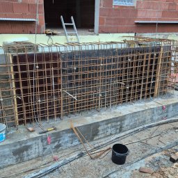 Jakub Cyrwus Firma Handlowo Usługowa - Zbrojenie fundamentu z deskowaniem i wystającymi prętami, widoczne narzędzia i materiały budowlane. Etap prac fundamentowych przed zalaniem betonem.