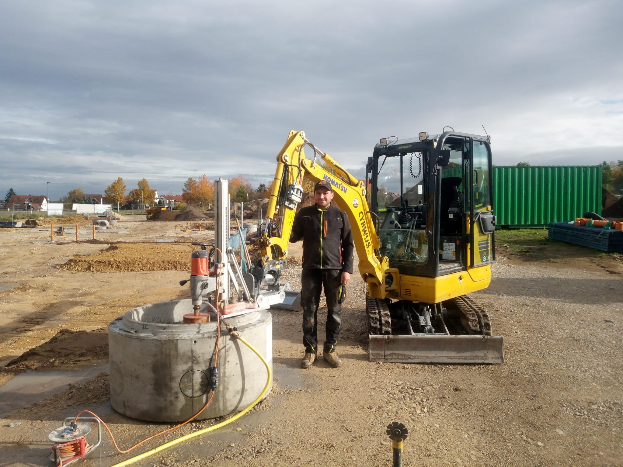 Pracownik z wiertnicą diamentową wierci otwór w betonowym kręgu na placu budowy, obok stoi żółta mini koparka Komatsu, w tle widoczny teren budowy i kontener.