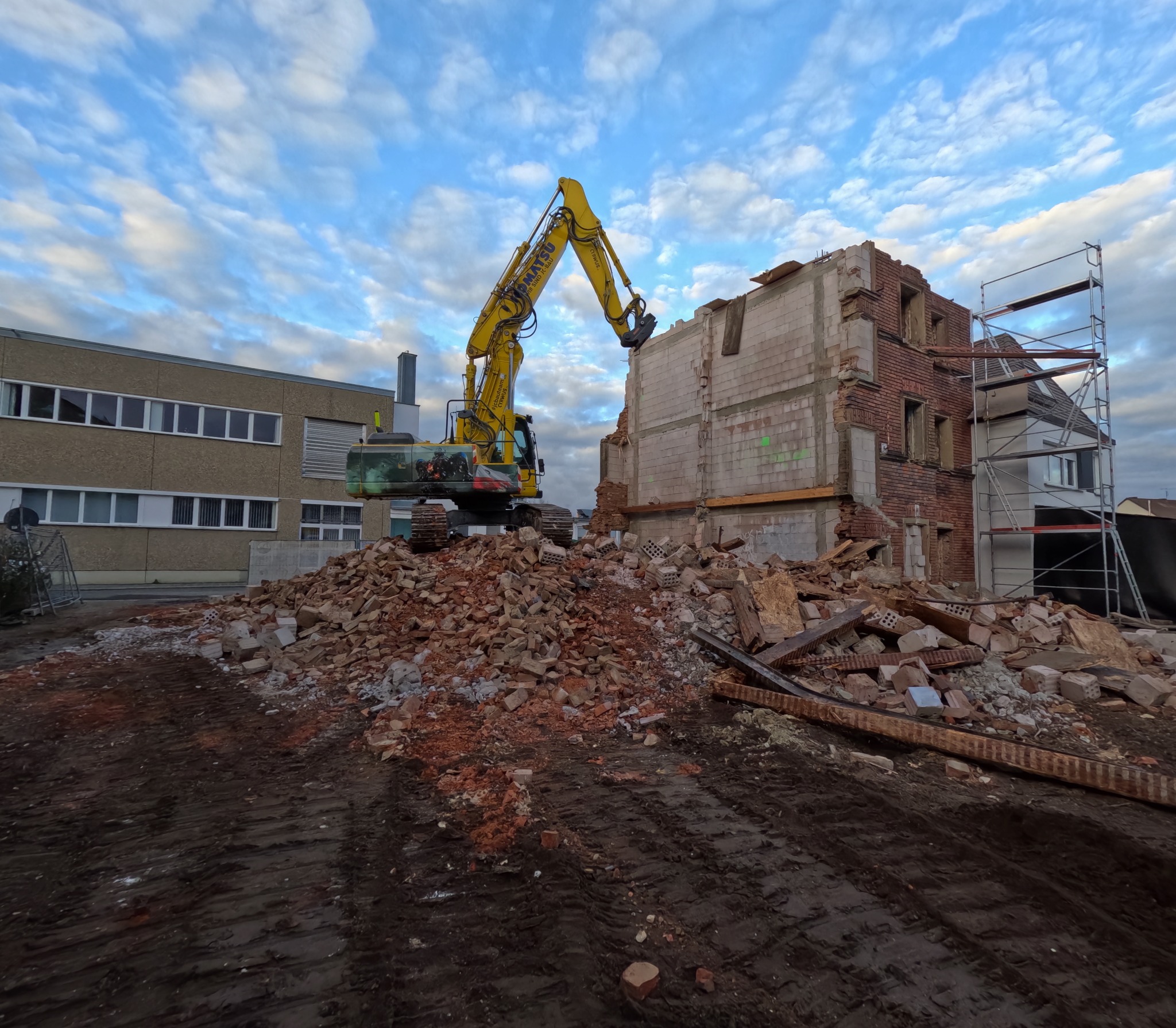 Żółta koparka na gruzowisku podczas rozbiórki budynku z cegły i pustaków. W tle budynek biurowy i rusztowanie. Błękitne niebo z chmurami.