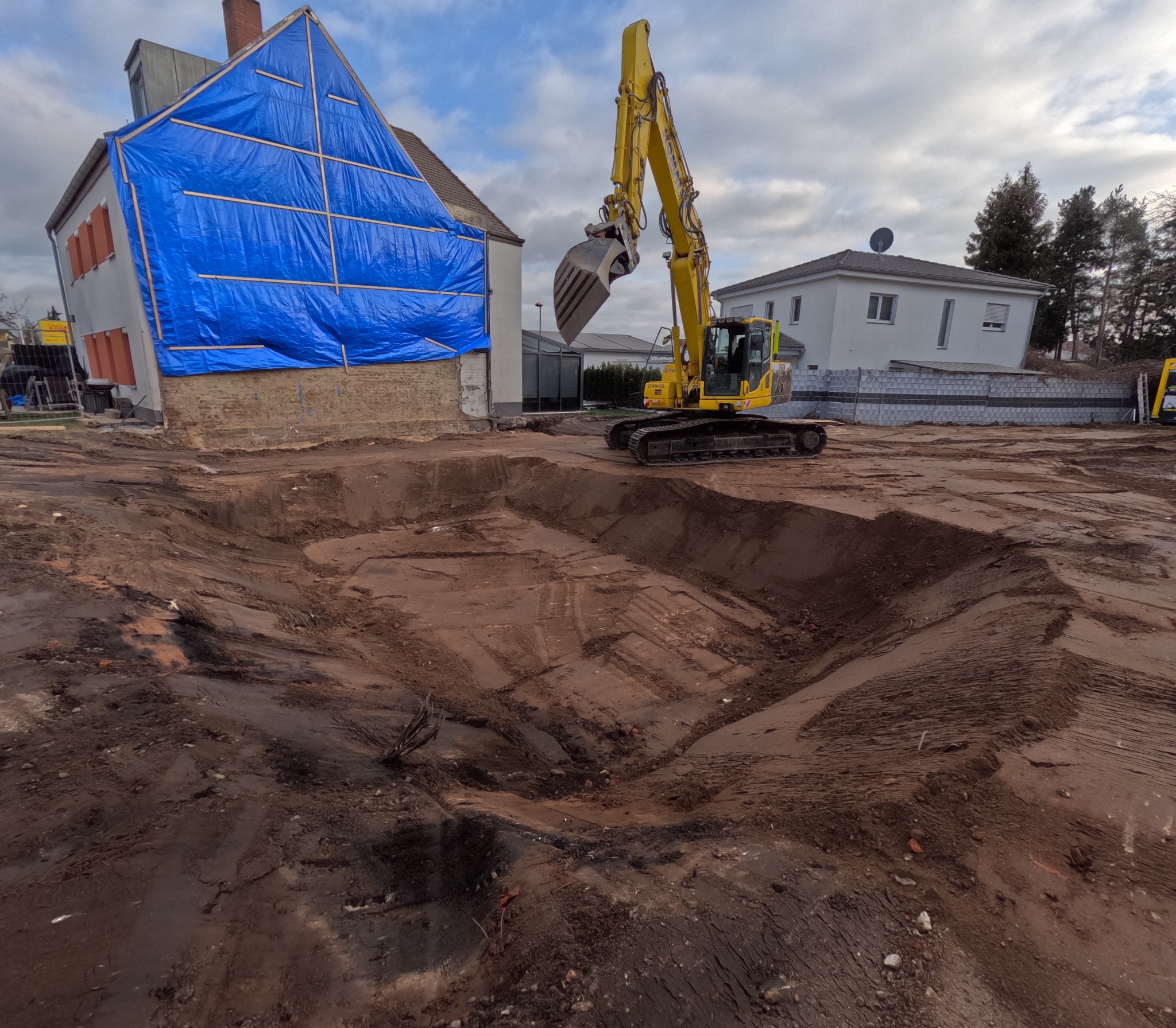 Żółta koparka na placu budowy, wykop pod fundamenty domu zabezpieczonego niebieską plandeką. Widoczne ślady opon i wykopana ziemia.
