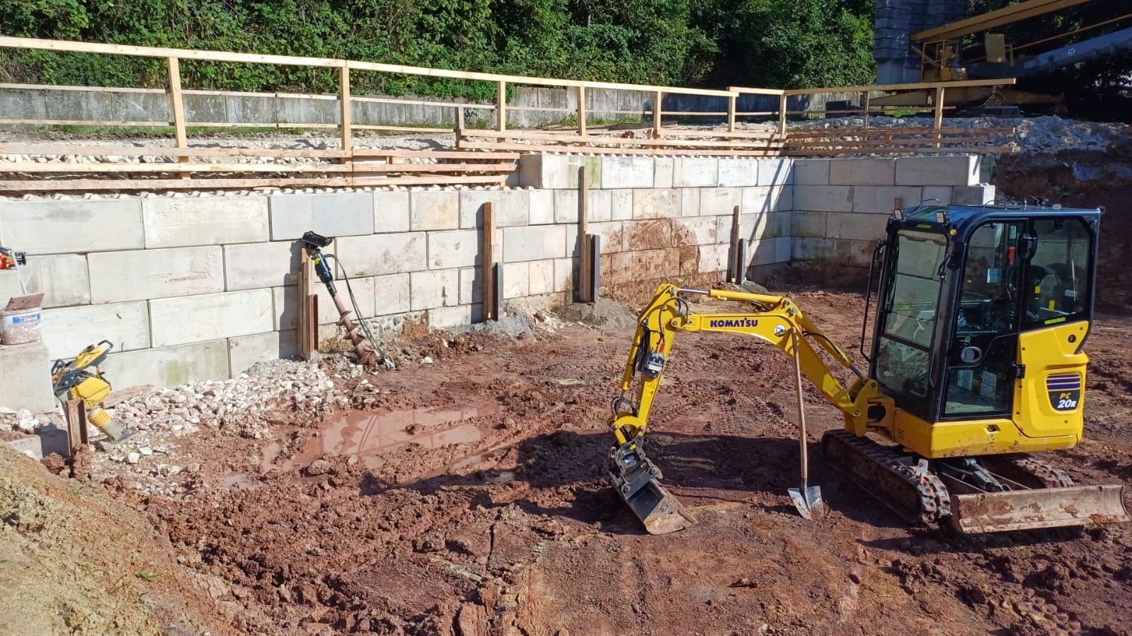 Żółta mini koparka Komatsu PC20R na placu budowy, obok betonowego muru oporowego z drewnianą balustradą. W tle zieleń drzew i fragment żurawia.