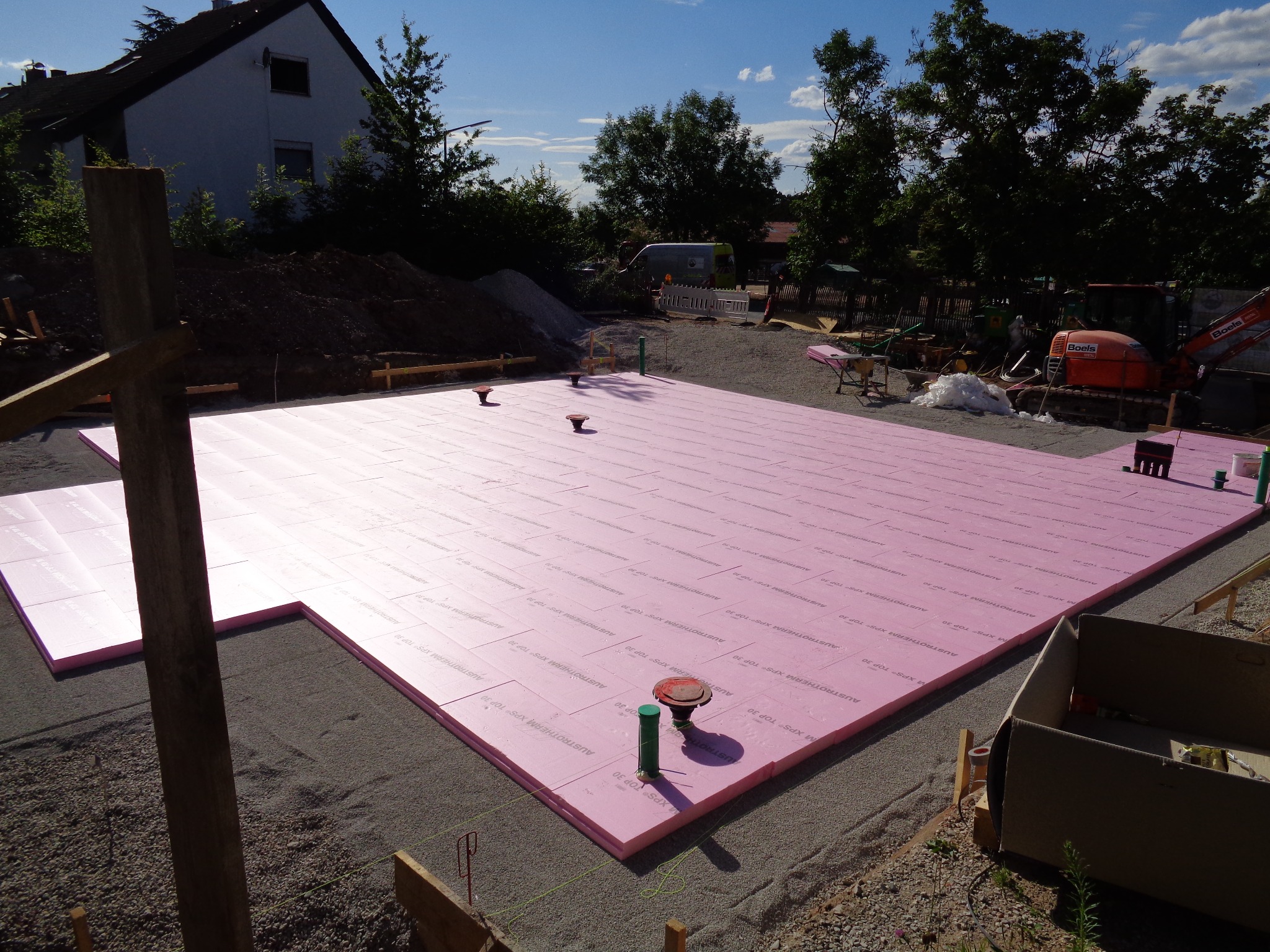 Różowa izolacja termiczna ułożona na przygotowanym podłożu pod fundamenty domu. Widoczne elementy instalacji, ziemia, drzewa i budynek w tle.