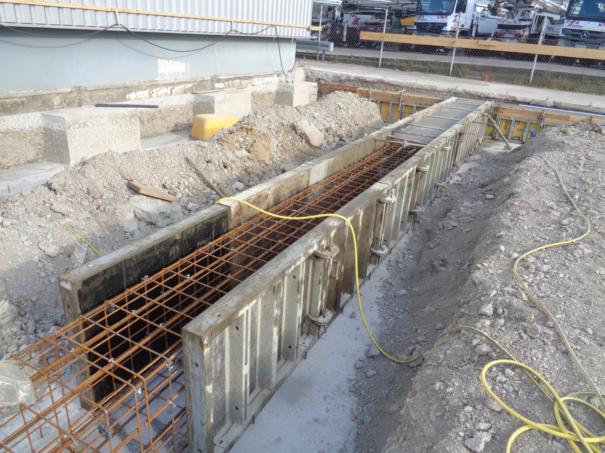 Zbrojenie w szalunku, przygotowane do zalania betonem. Widoczne metalowe pręty tworzące siatkę i żółty kabel. W tle budynek i betoniarki.