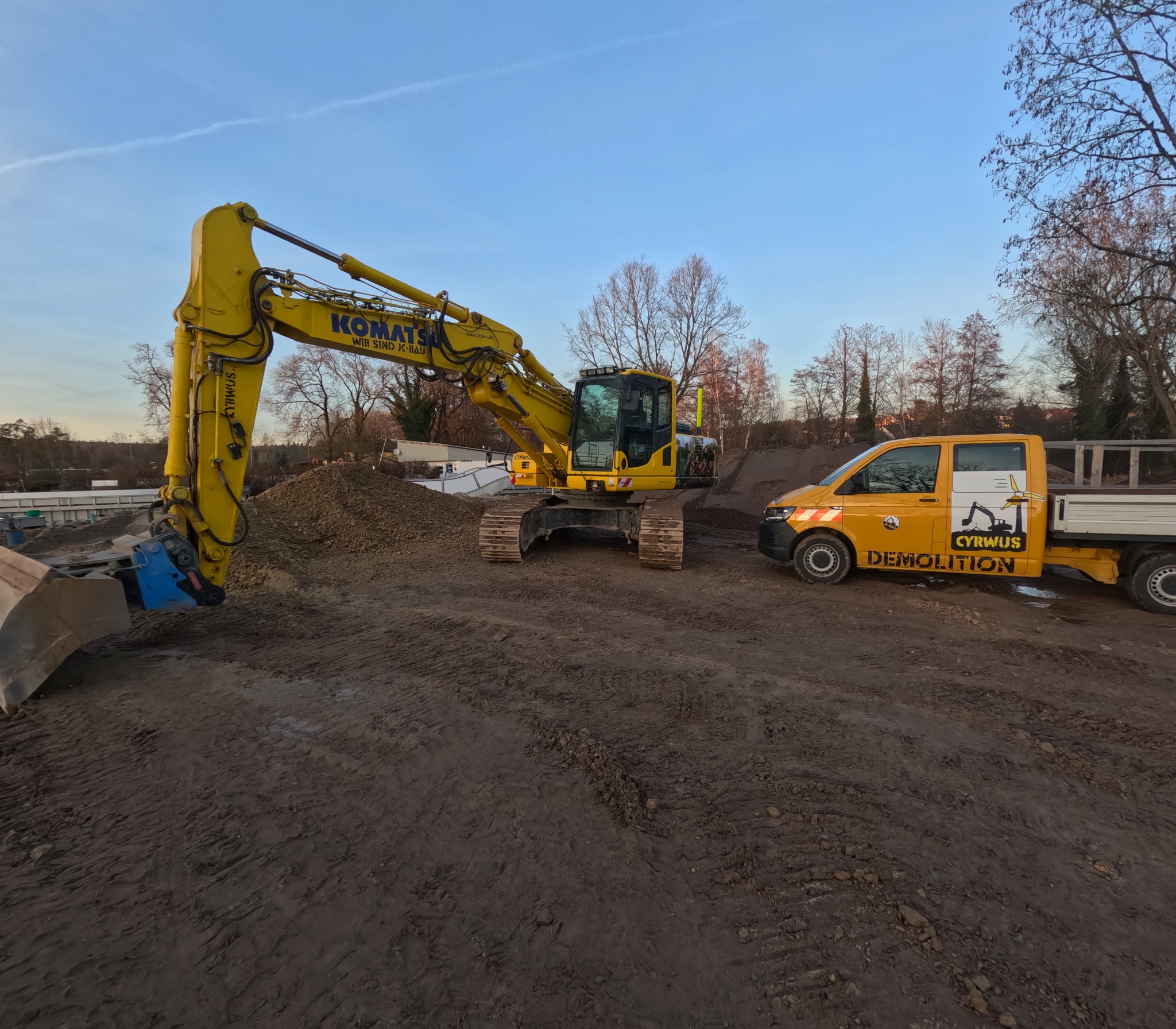 Żółta koparka Komatsu z młotem hydraulicznym i żółty samochód z napisem DEMOLITION na placu budowy. W tle drzewa i błękitne niebo.