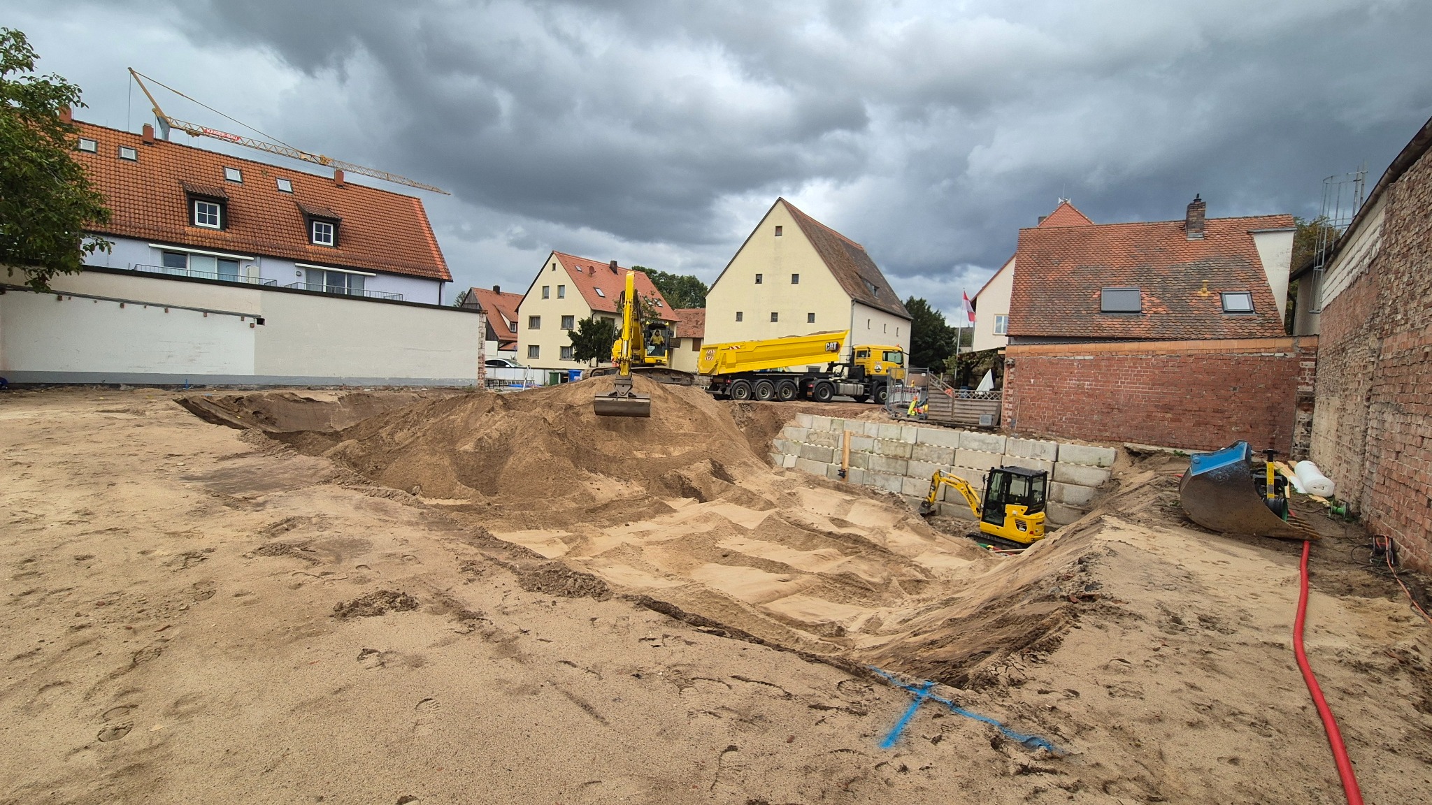 Głęboki wykop pod budowę z żółtym sprzętem ciężkim (koparka i wywrotka) na tle zabudowy miejskiej i pochmurnego nieba. Widoczne betonowe bloki oporowe.