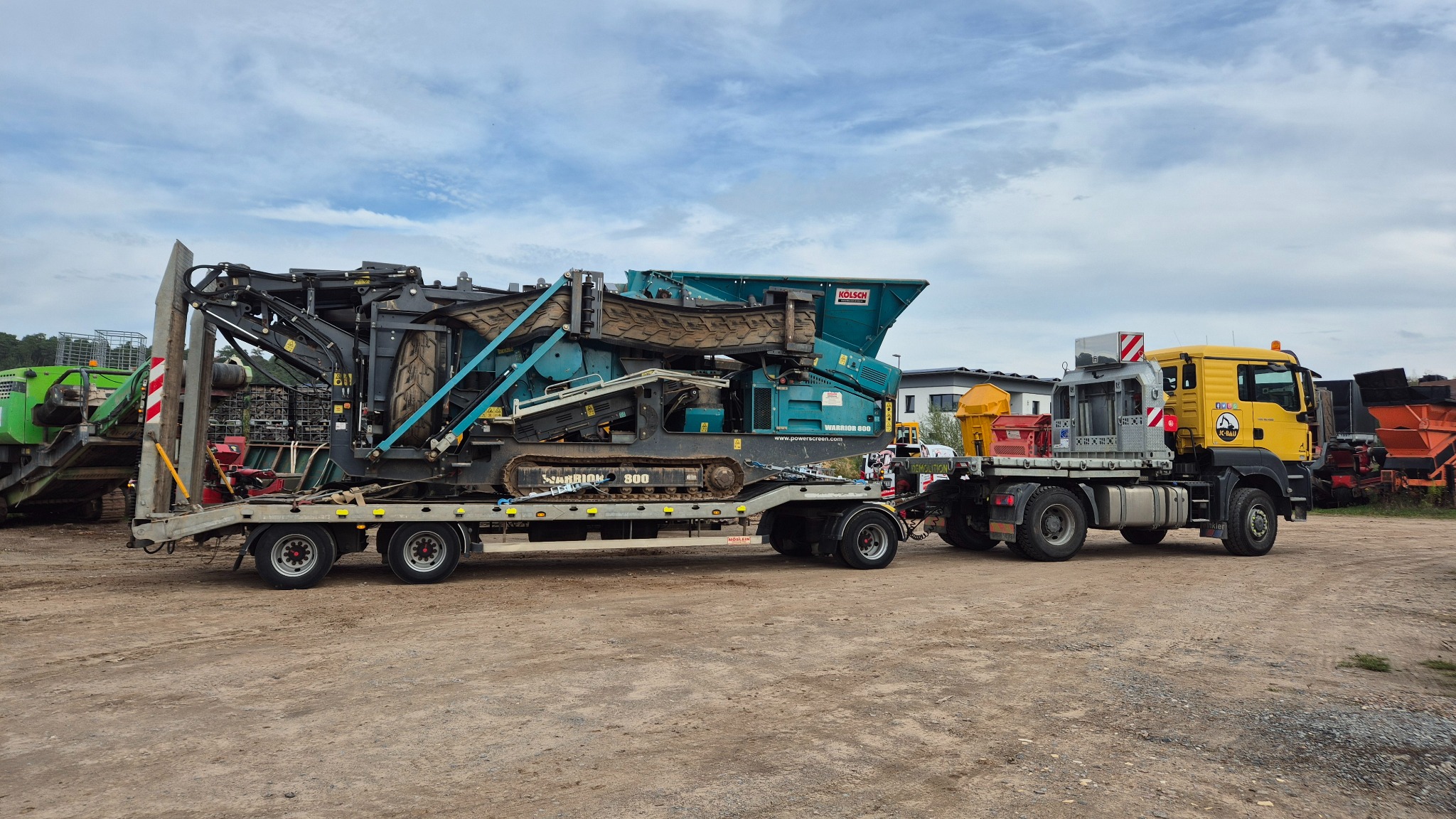 Transport kruszarki Warrior 800 na lawecie przez żółtą ciężarówkę JC-Bau w Waksmundzie. Sprzęt do robót ziemnych na placu budowy, widok z boku, pochmurne niebo.