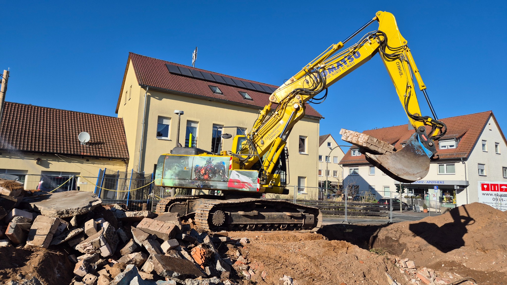Żółta koparka Komatsu z grafiką na tle budynku podczas prac rozbiórkowych. W łyżce gruz, w tle budynki mieszkalne i błękitne niebo.