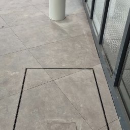 Michał Murdza - Szara, kwadratowa posadzka przemysłowa z odpływem liniowym, ułożona w regularny wzór. Widoczny fragment kolumny i okna. Jasne, naturalne światło.