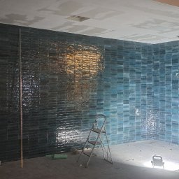 Michał Murdza - Ściany wyłożone błyszczącą, turkusową cegiełką w trakcie remontu łazienki. Widoczna drabina, lampa budowlana i kable. Częściowo położona glazura.