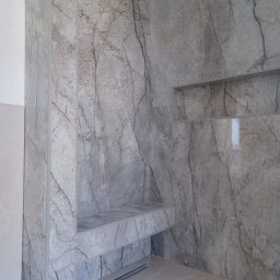 Michał Murdza - Wykończona łazienka z szarymi płytkami imitującymi marmur na ścianach, wnęką i półką. Widoczny odpływ liniowy w podłodze. Minimalistyczny design.