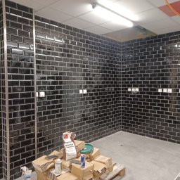 Michał Murdza - Czarne, błyszczące płytki ułożone w cegiełkę na ścianach. Widoczne puszki elektryczne, materiały budowlane na palecie i fragment sufitu z oświetleniem.