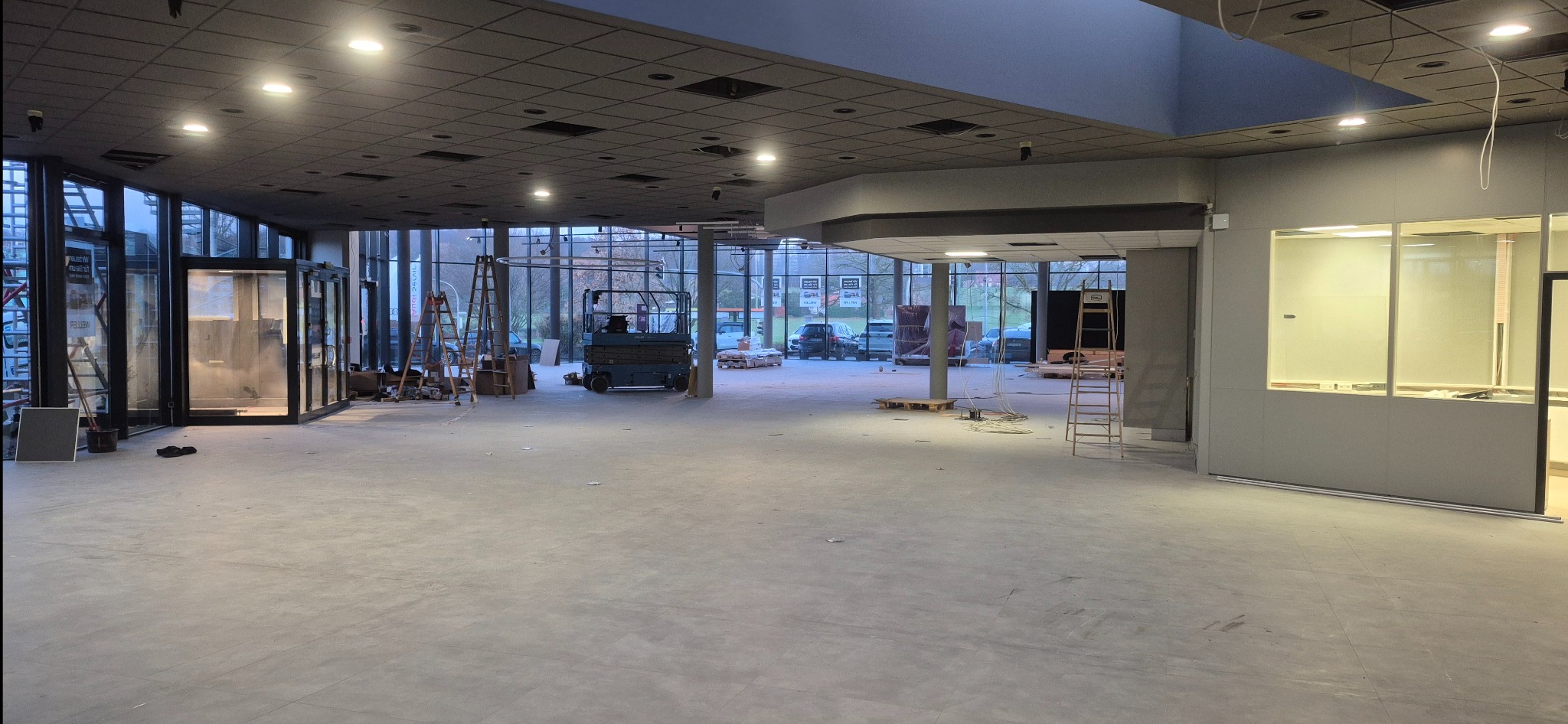 Wnętrze komercyjne w trakcie prac wykończeniowych: nowa posadzka, drabiny, podnośnik nożycowy, duże okna. Widoczne przygotowania do dalszych prac.