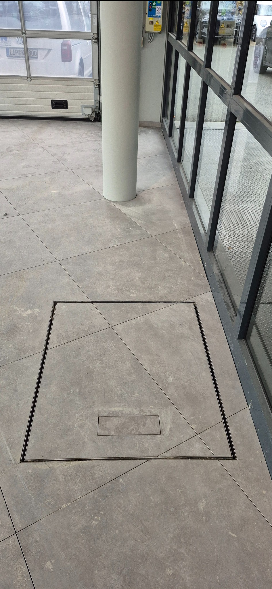 Szara, kwadratowa posadzka przemysłowa z odpływem liniowym, ułożona w regularny wzór. Widoczny fragment kolumny i okna. Jasne, naturalne światło.