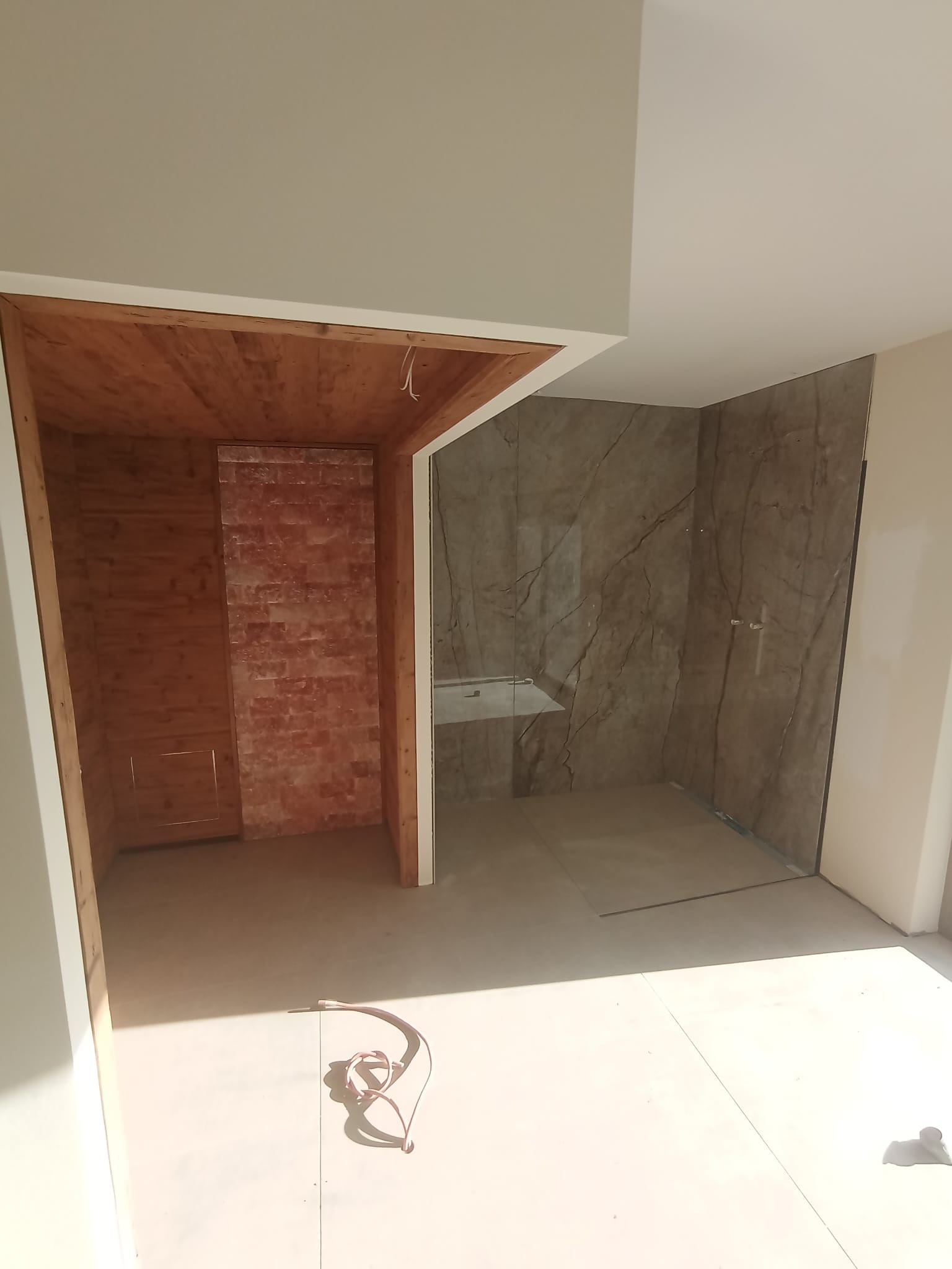 Łazienka w trakcie remontu: kabina prysznicowa z marmurowymi płytami, obok drewniana konstrukcja sauny w surowym stanie. Widoczne przewody na podłodze.