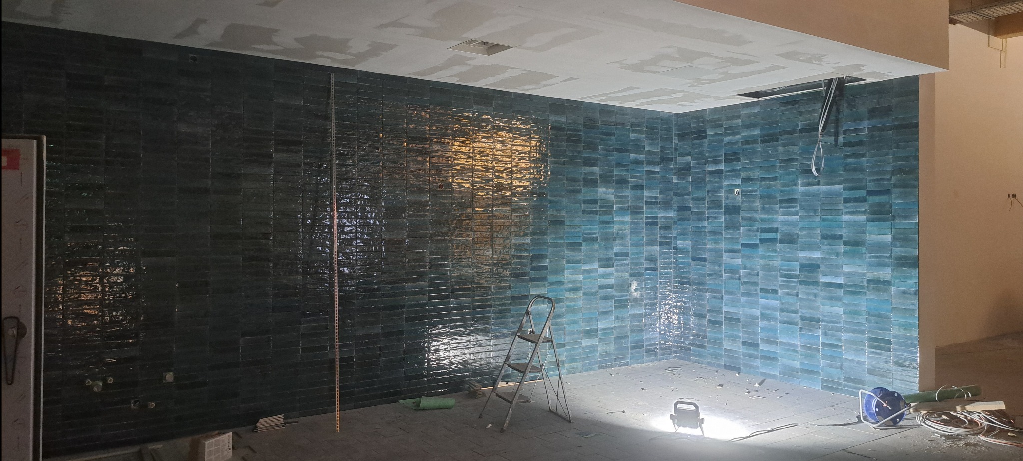 Ściany wyłożone błyszczącą, turkusową cegiełką w trakcie remontu łazienki. Widoczna drabina, lampa budowlana i kable. Częściowo położona glazura.