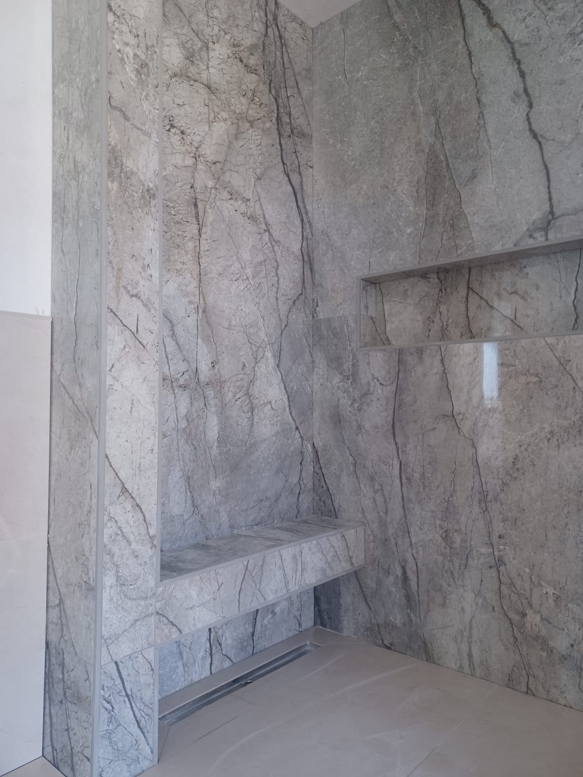 Wykończona łazienka z szarymi płytkami imitującymi marmur na ścianach, wnęką i półką. Widoczny odpływ liniowy w podłodze. Minimalistyczny design.