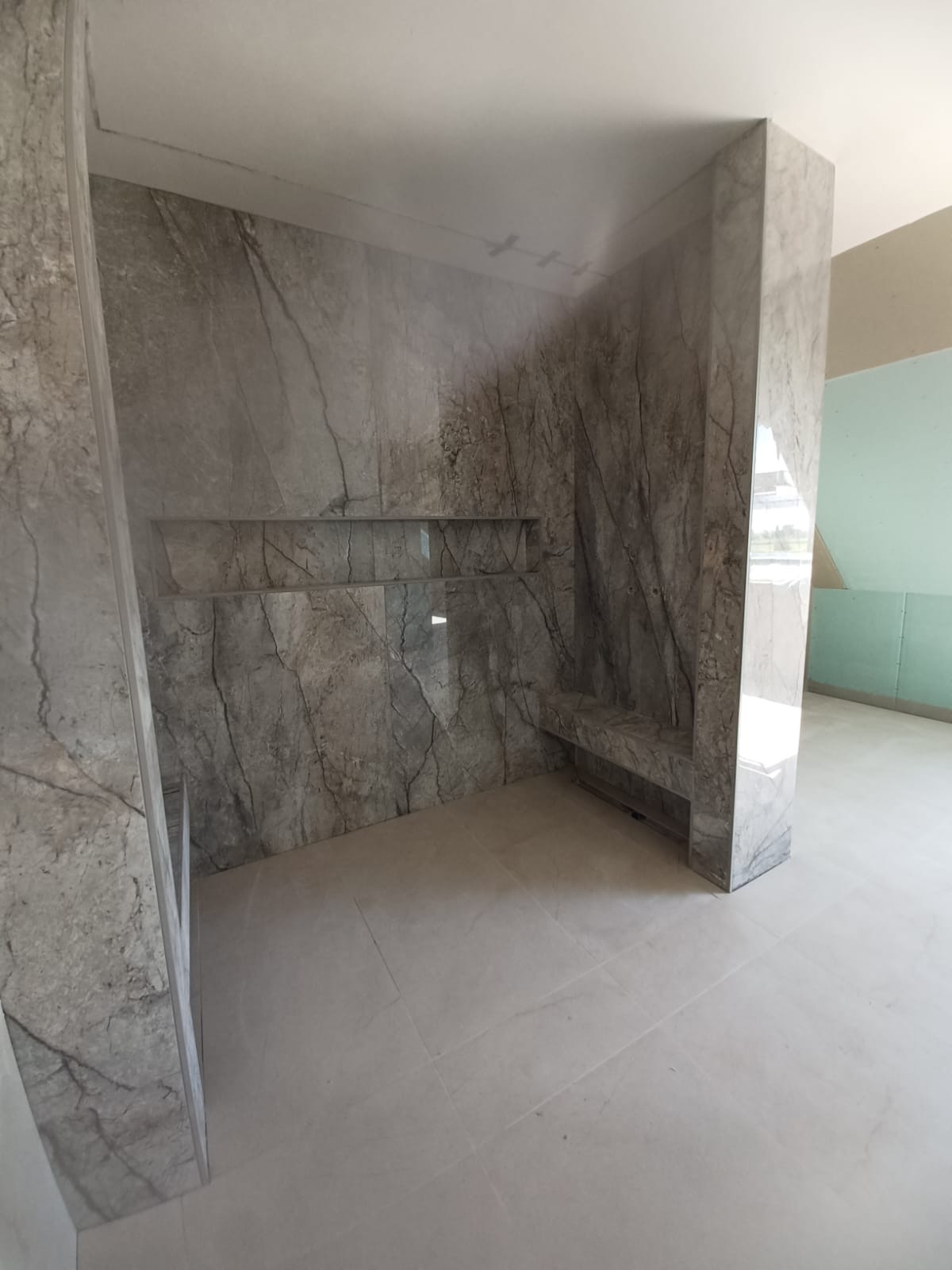 Wykończona łazienka z szarymi płytkami imitującymi marmur na ścianach i podłodze. W ścianie wnęki i siedzisko. Widok na sąsiednie pomieszczenie w trakcie prac.